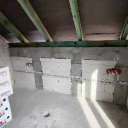 Wewnętrzne okablowanie elektryczne w surowym pomieszczeniu, widoczne puszki instalacyjne i przewody w białych listwach, ściany z betonu komórkowego, drewniany szkielet stropu.