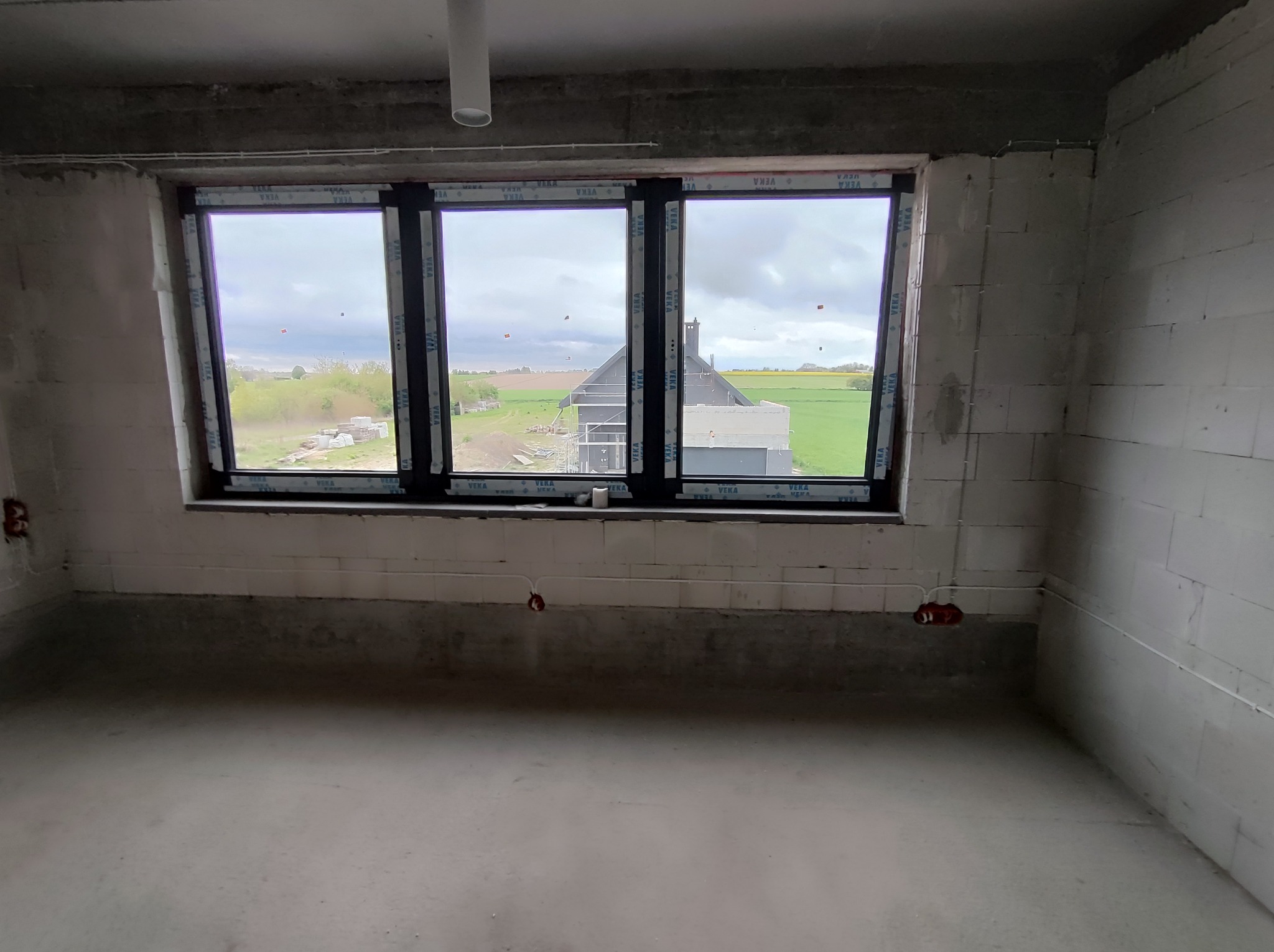 Surowe wnętrze w stanie deweloperskim z trzema oknami PCV marki VEKA, widok na budowę domu i zielony krajobraz za oknem, widoczne wystające kable elektryczne.