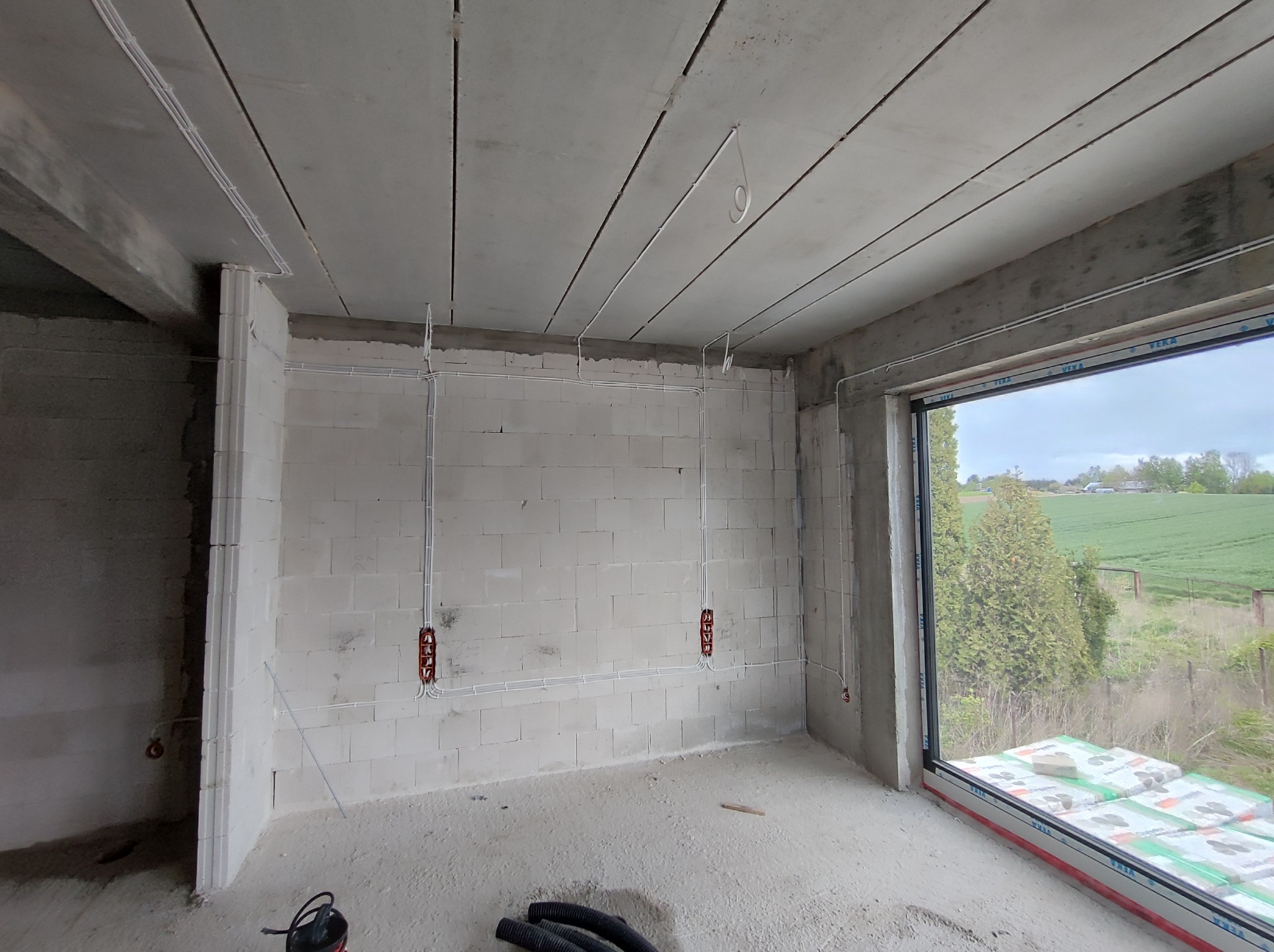 Surowy stan wnętrza z widoczną instalacją elektryczną w peszlach na ścianach z betonu komórkowego oraz pod sufitem z płyt prefabrykowanych. Widoczne puszki elektryczne i przewody. W tle widok...