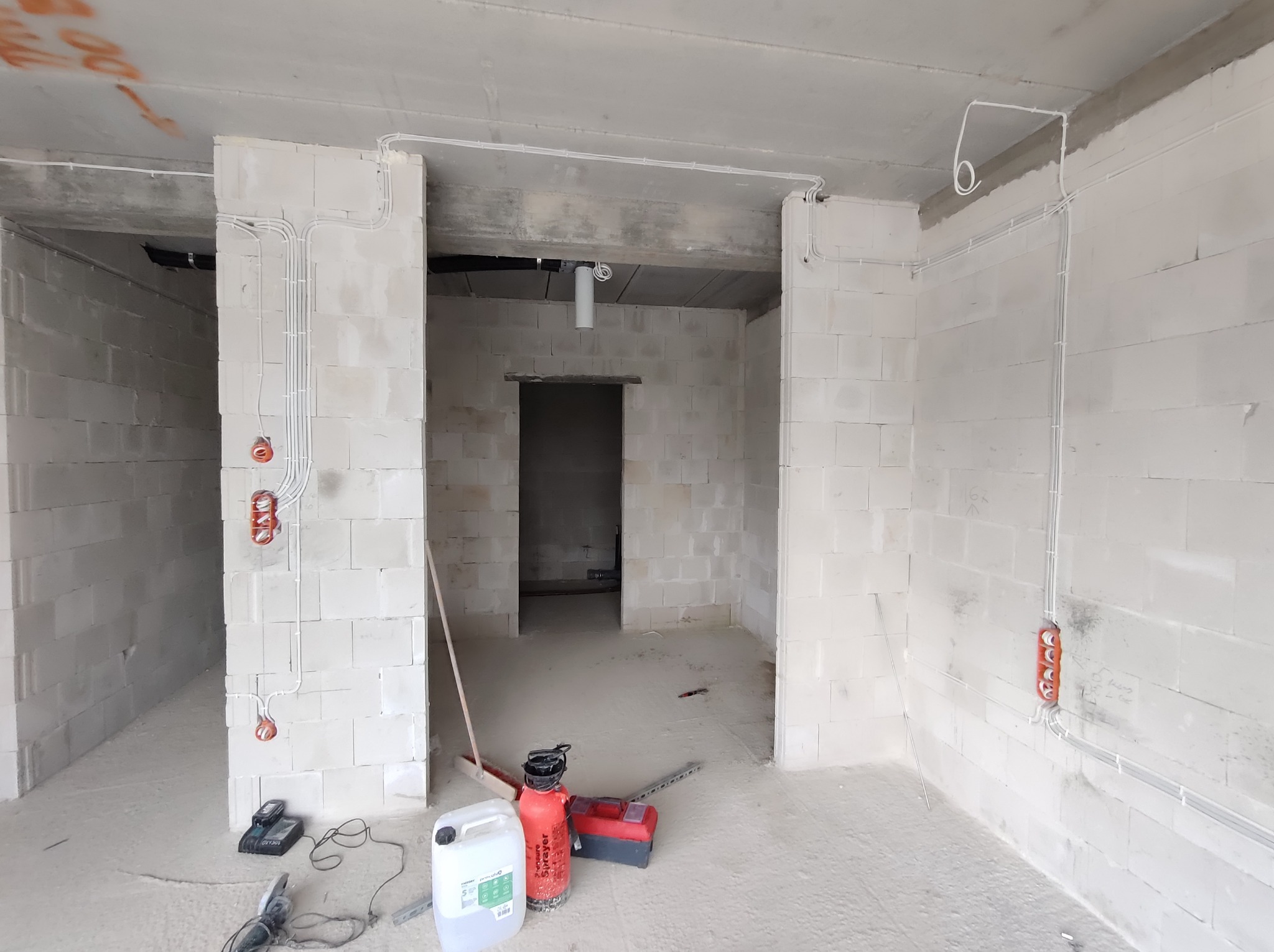 Surowy stan wnętrza budynku w trakcie prac elektrycznych: widoczne białe ściany z betonu komórkowego, rozprowadzone białe kable elektryczne w peszlach, puszki instalacyjne, narzędzia i pojemnik...