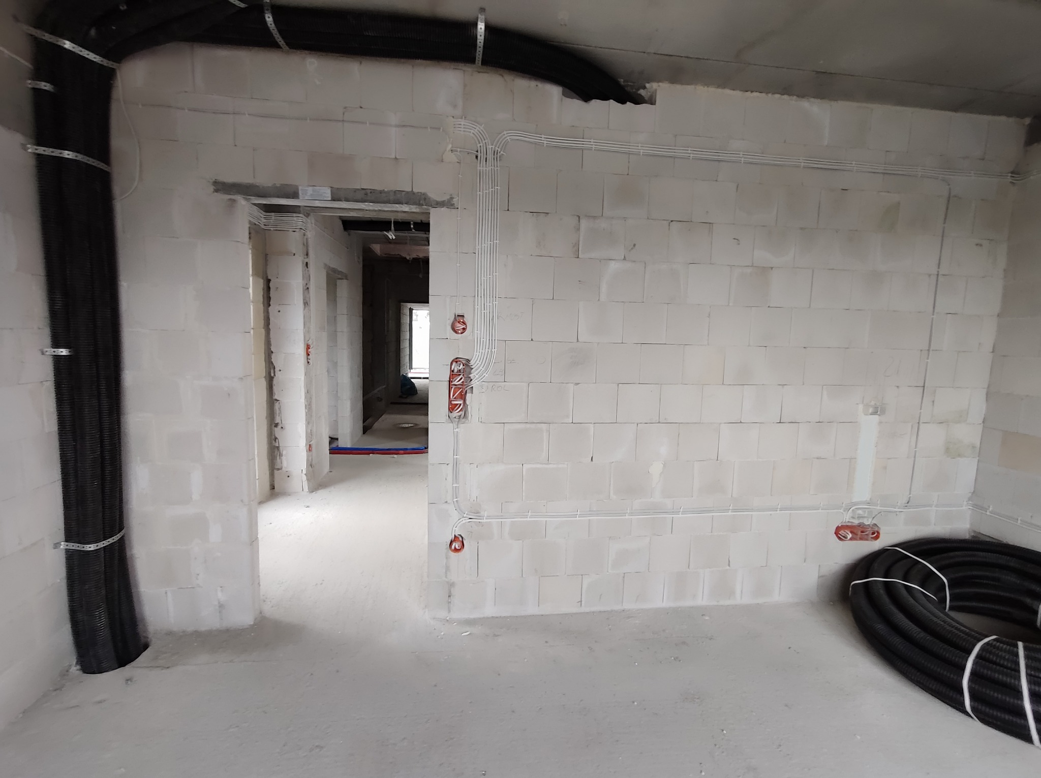 Surowy stan wykończenia wnętrza z widocznymi przewodami elektrycznymi prowadzonymi po ścianach z betonu komórkowego oraz zwiniętym w krąg peszlem na podłodze.