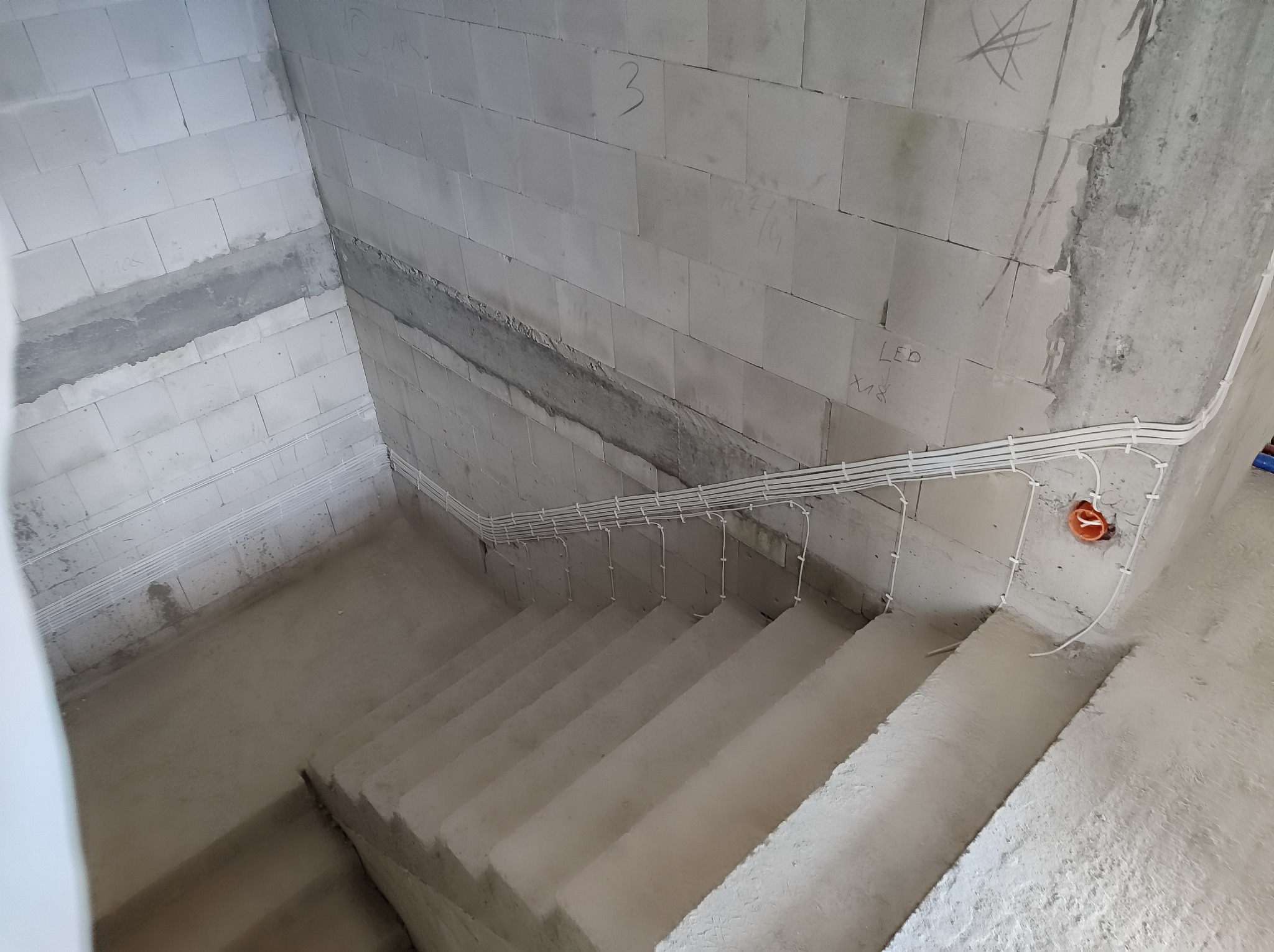 Surowe schody w trakcie budowy z widocznymi przewodami elektrycznymi przymocowanymi do ściany z betonu komórkowego, z pomarańczową puszką instalacyjną.