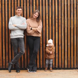 Sesje rodzinne, portretowe, pertnerskie - Rodzinne zdjęcie: młoda para z dzieckiem w jesiennych ubraniach, pozująca na tle drewnianej ściany z pionowych desek.
