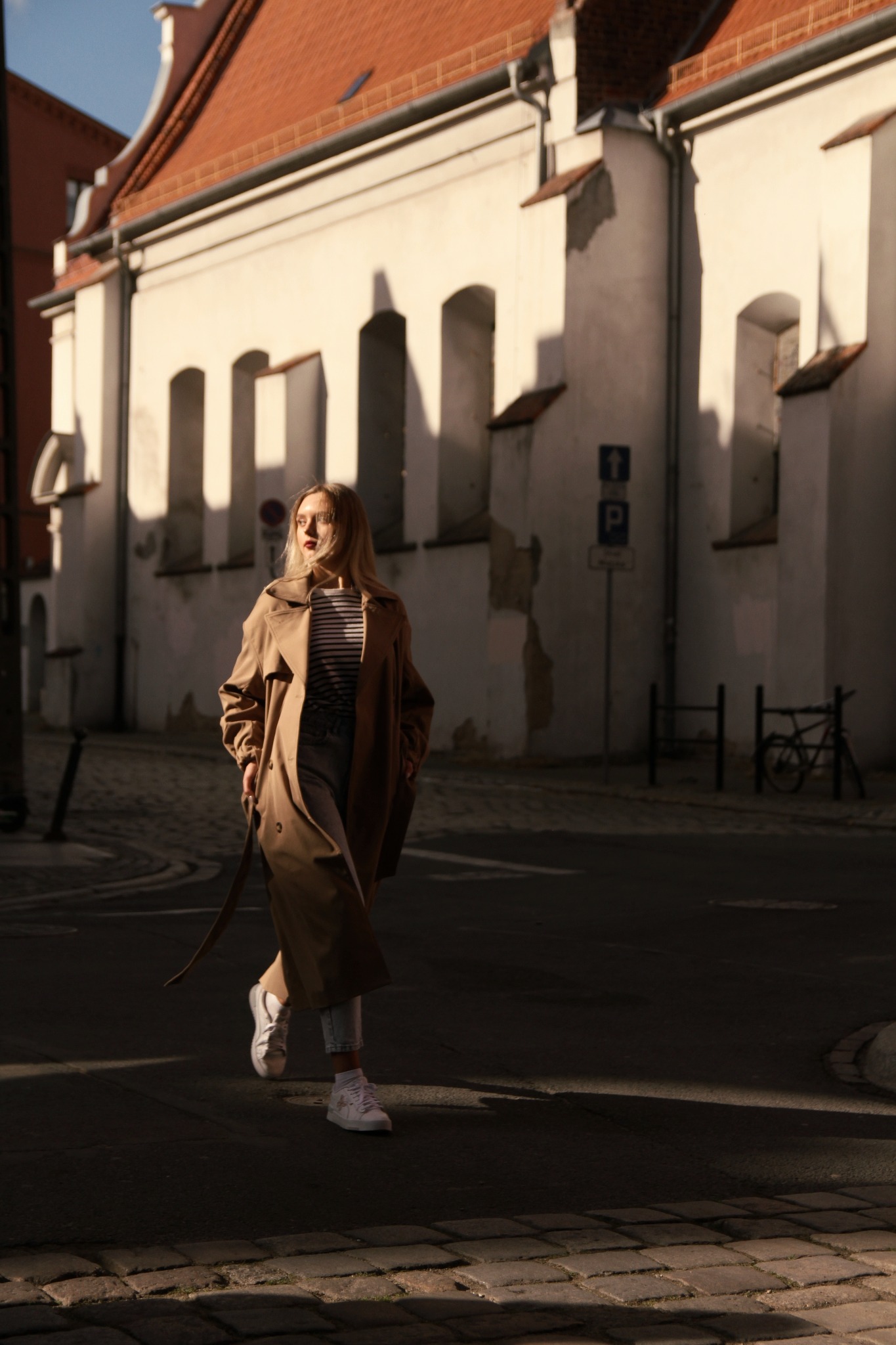 Kobieta w beżowym płaszczu przechodzi przez zacienioną ulicę, w tle budynek z czerwonym dachem i białą elewacją z arkadowymi oknami.