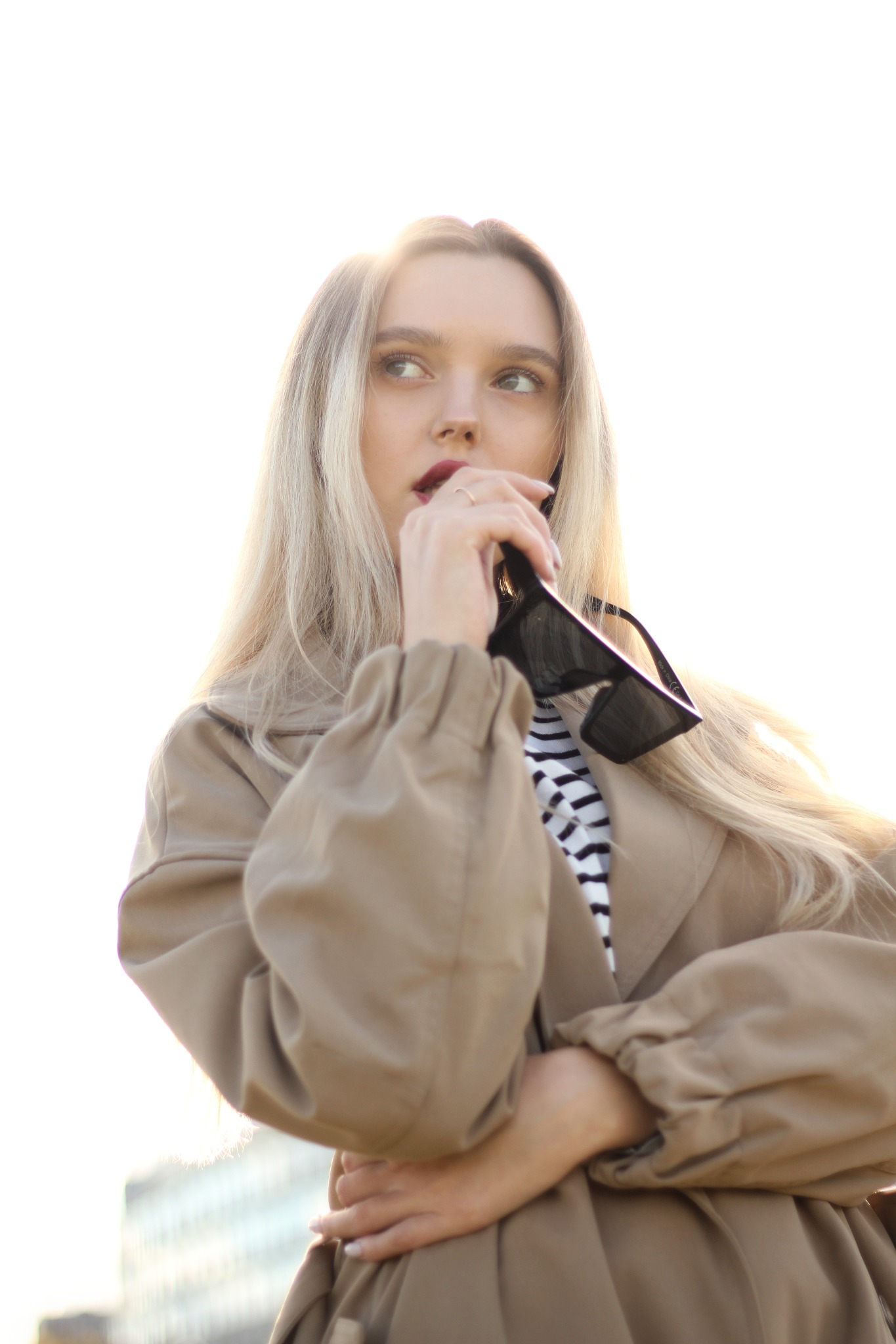 Portret kobiety z blond włosami w beżowym płaszczu, trzymającej okulary przeciwsłoneczne przy ustach, spoglądającej w górę na tle rozmytego budynku.