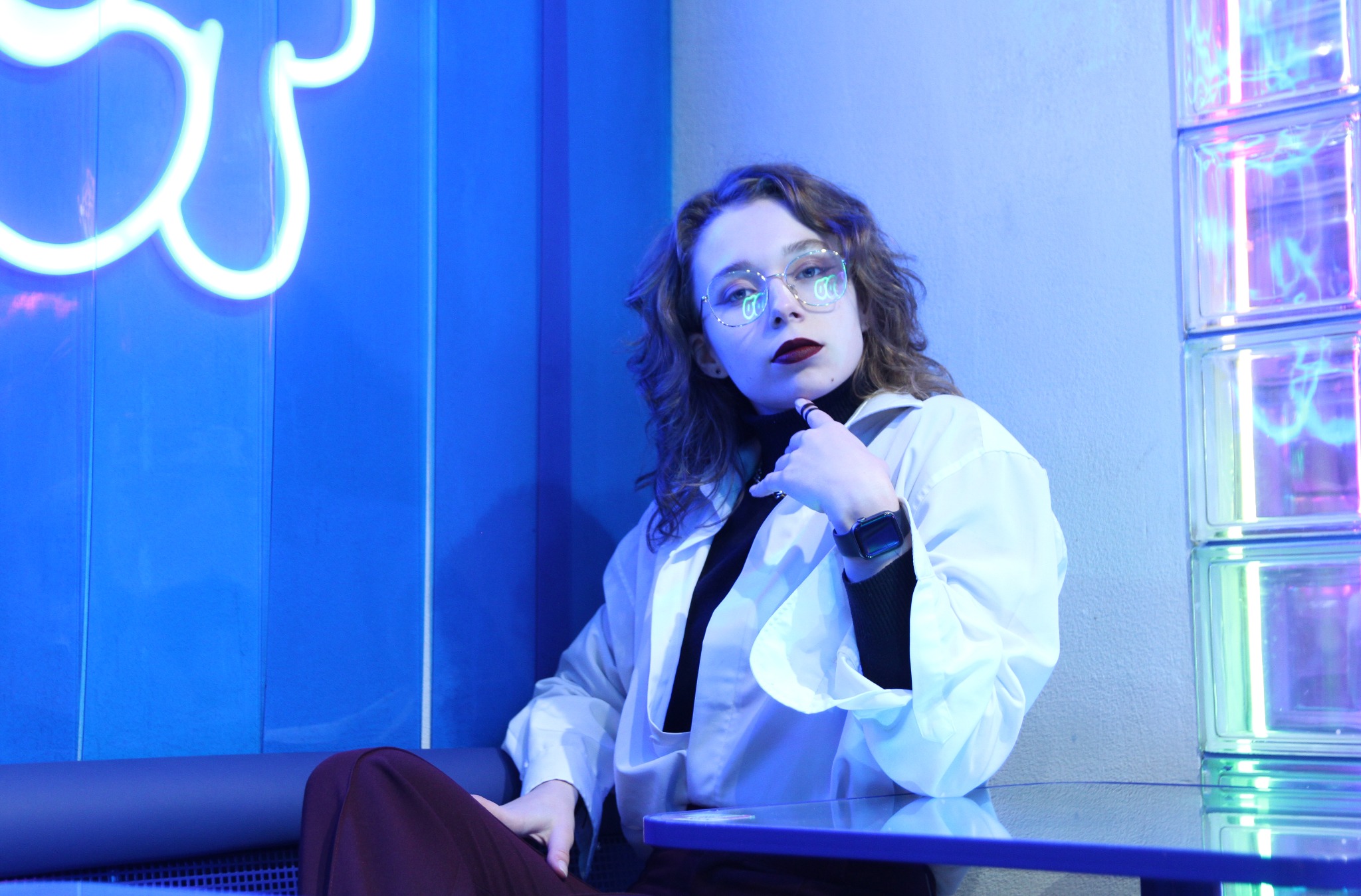Portret kobiety w okularach, z ciemną szminką, pozującej w pomieszczeniu z neonowym znakiem w kształcie chmury i dekoracyjnymi luksferami, oświetlonym intensywnym niebieskim światłem.