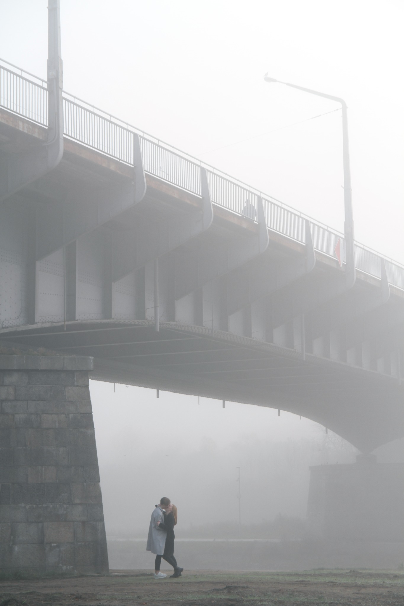 Para całująca się w mglisty poranek pod mostem, delikatne światło podkreśla romantyczny nastrój, w tle widoczna latarnia.