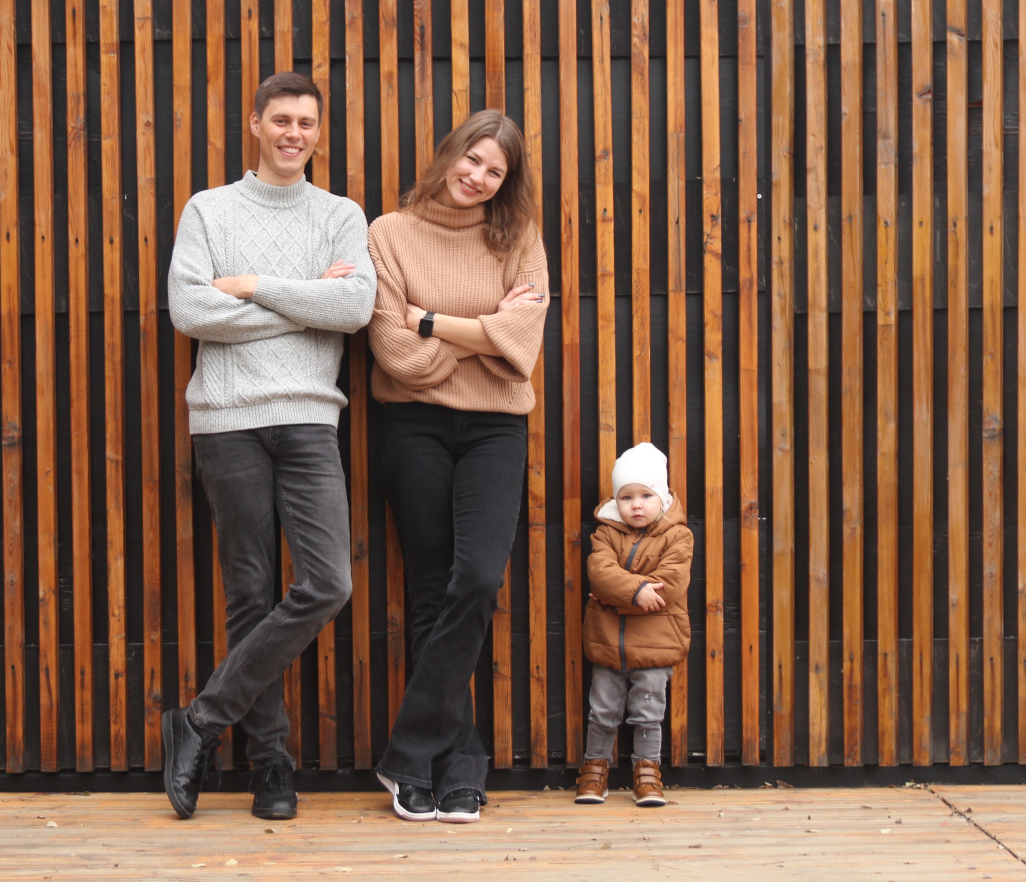 Rodzinne zdjęcie: młoda para z dzieckiem w jesiennych ubraniach, pozująca na tle drewnianej ściany z pionowych desek.