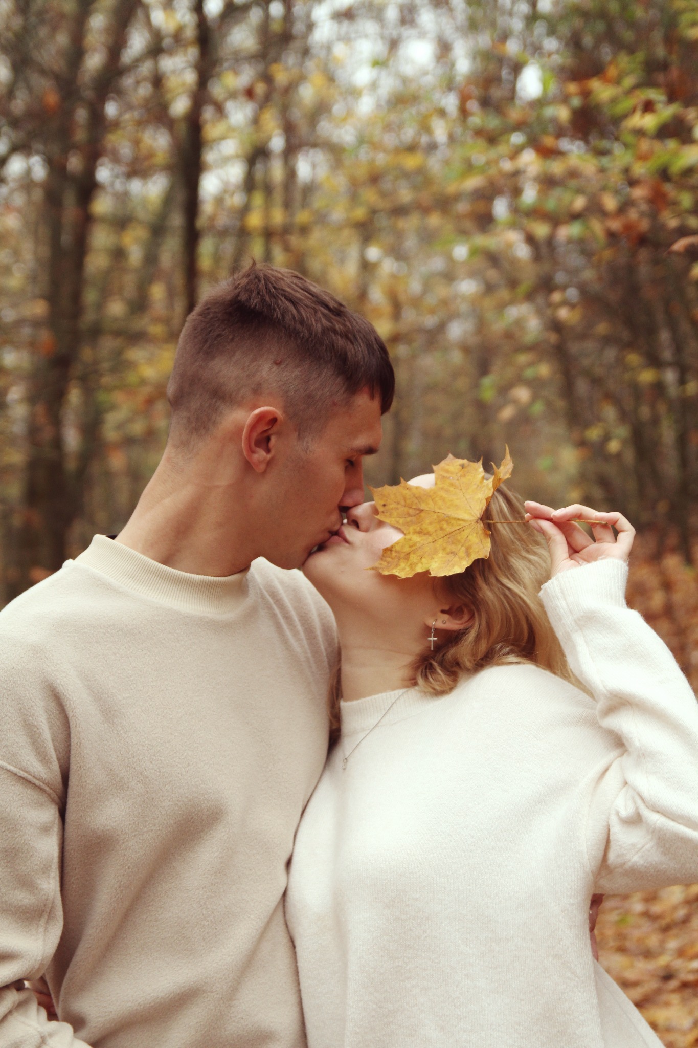 Zakochana para całuje się w jesiennym lesie, dziewczyna trzyma żółty liść na twarzy, widoczne drzewa z opadającymi liśćmi w tle.
