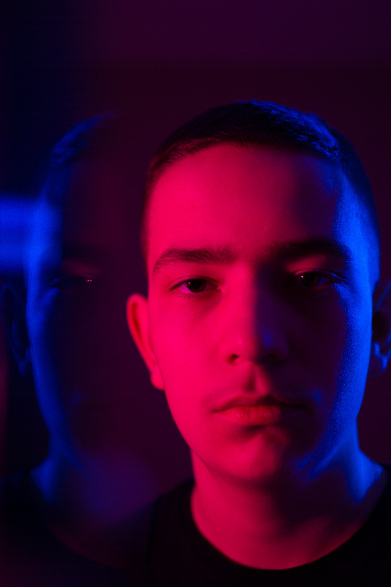 Portret młodego mężczyzny oświetlonego czerwonym i niebieskim światłem, z delikatnym echem jego sylwetki w tle.