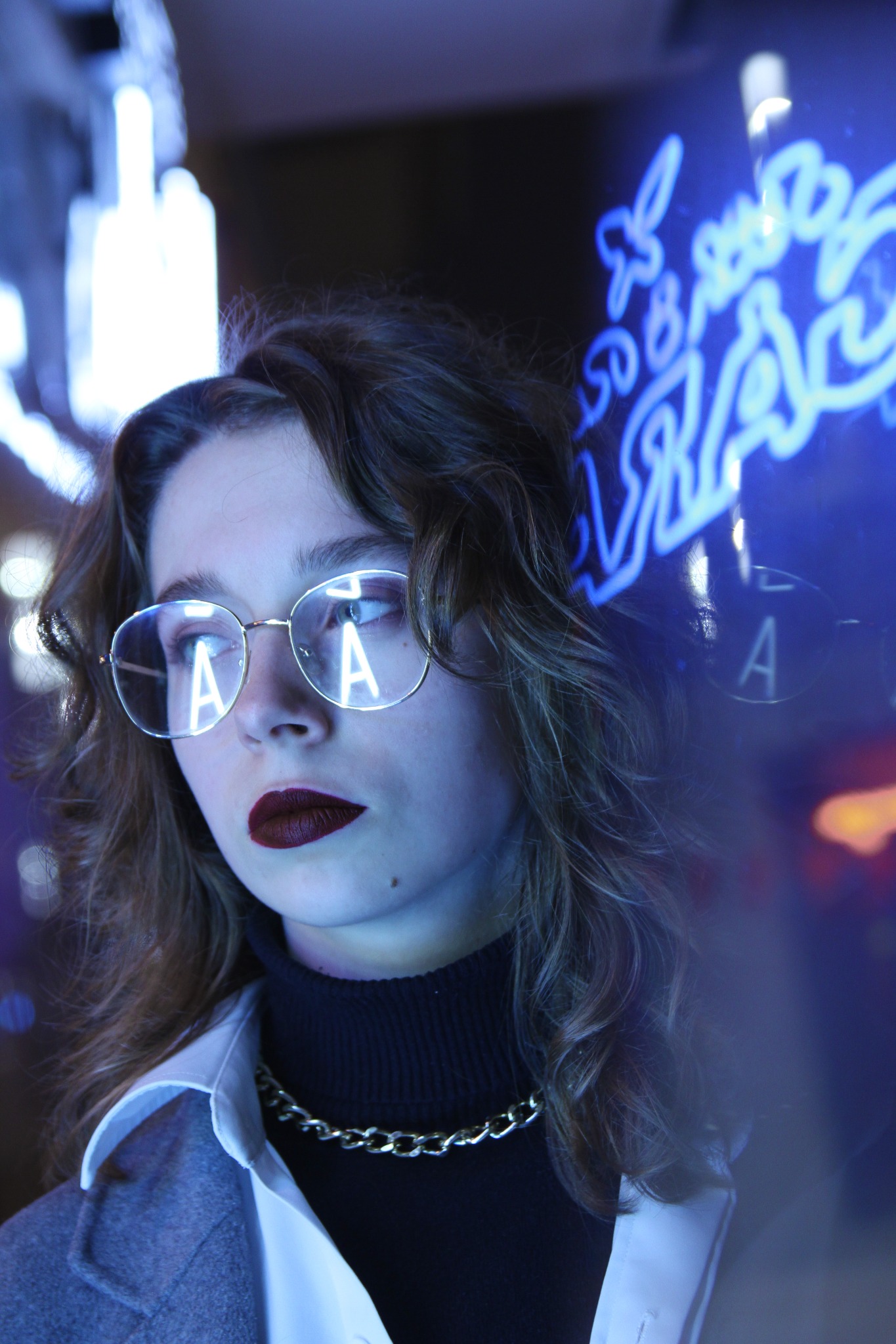 Portret kobiety w okularach z odbiciem neonu, ciemna szminka, czarny golf i złoty łańcuch na tle niebieskiego neonu.