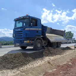 Niebieska ciężarówka Scania wyładowuje gruz na poboczu drogi, w tle widoczne góry i błękitne niebo z chmurami.