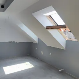 Poddasze po remoncie z szarymi ścianami, dwoma oknami dachowymi i ciemnymi drzwiami wejściowymi, widoczne niedociągnięcia w postaci niezakrytych przewodów elektrycznych i śladów po szpachlowaniu.