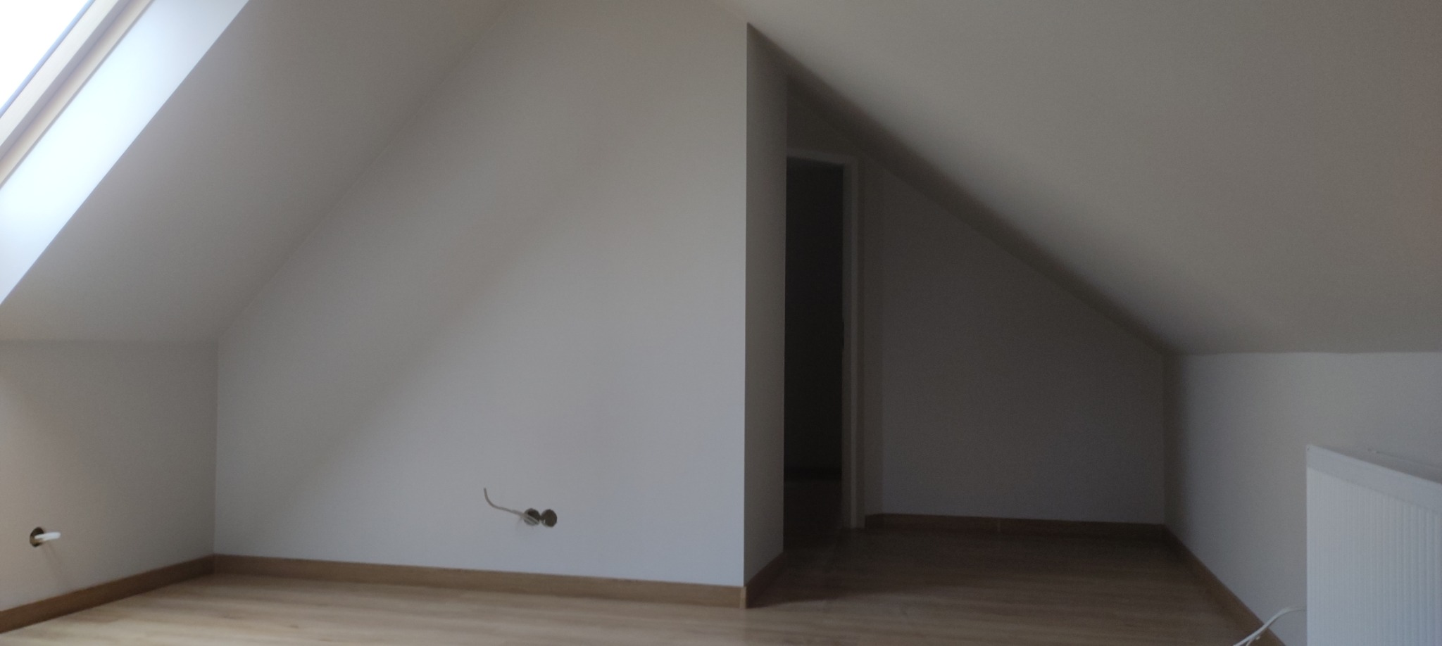 Poddasze po remoncie z oknem dachowym, widoczne gładkie białe ściany, drewniana podłoga i wystające kable elektryczne ze ściany.