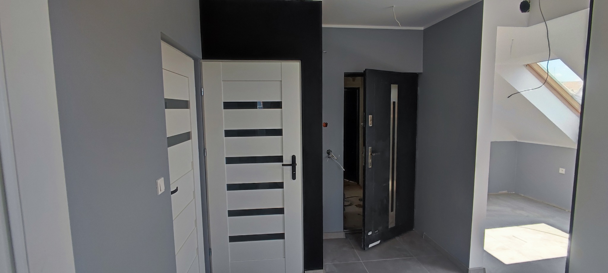 Wnętrze po odświeżeniu: szare ściany, białe drzwi wewnętrzne z poziomymi, szarymi paskami, otwarte, ciemne drzwi wejściowe z widocznymi kablami na ścianie i oknem dachowym w tle.