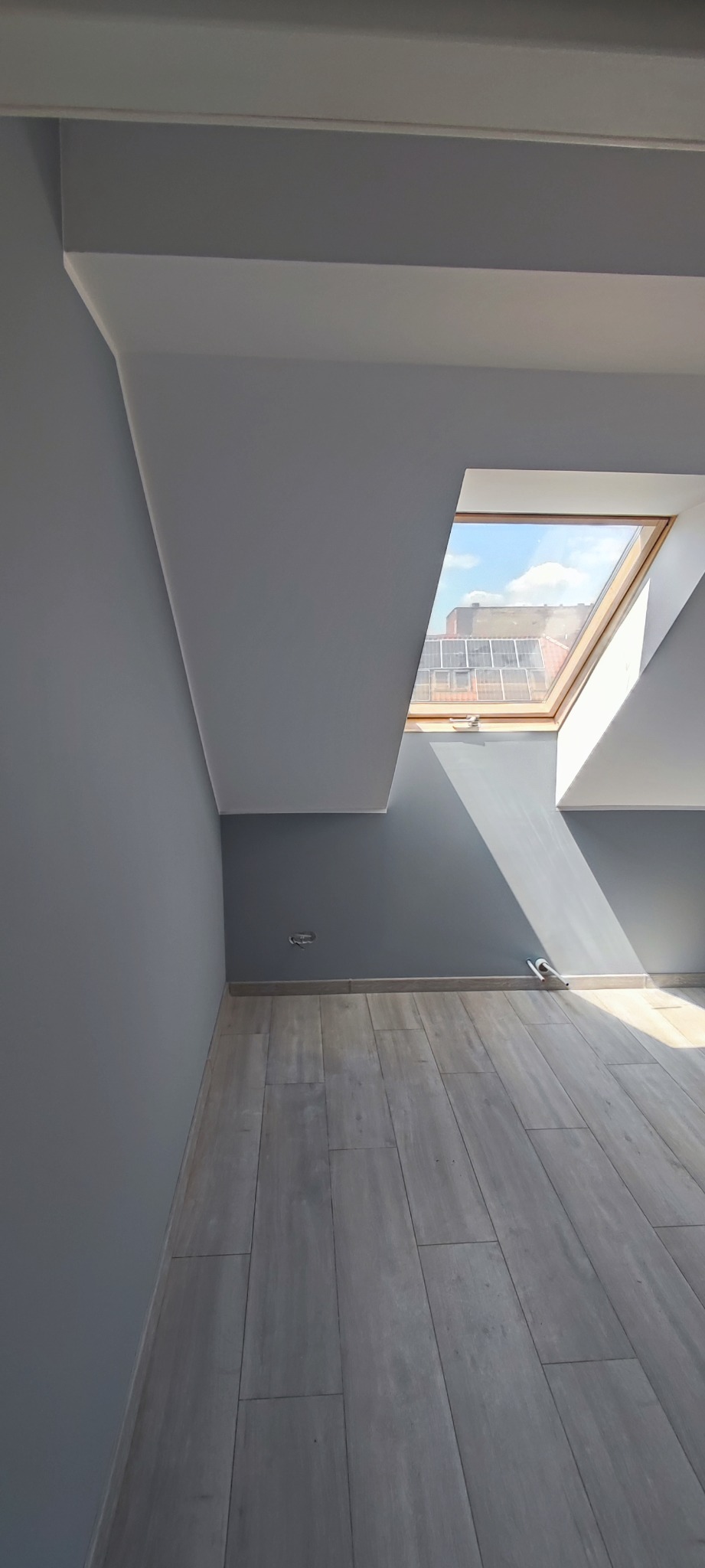 Poddasze po remoncie: szare ściany, drewnopodobne panele podłogowe i okno dachowe wpuszczające promienie słoneczne.