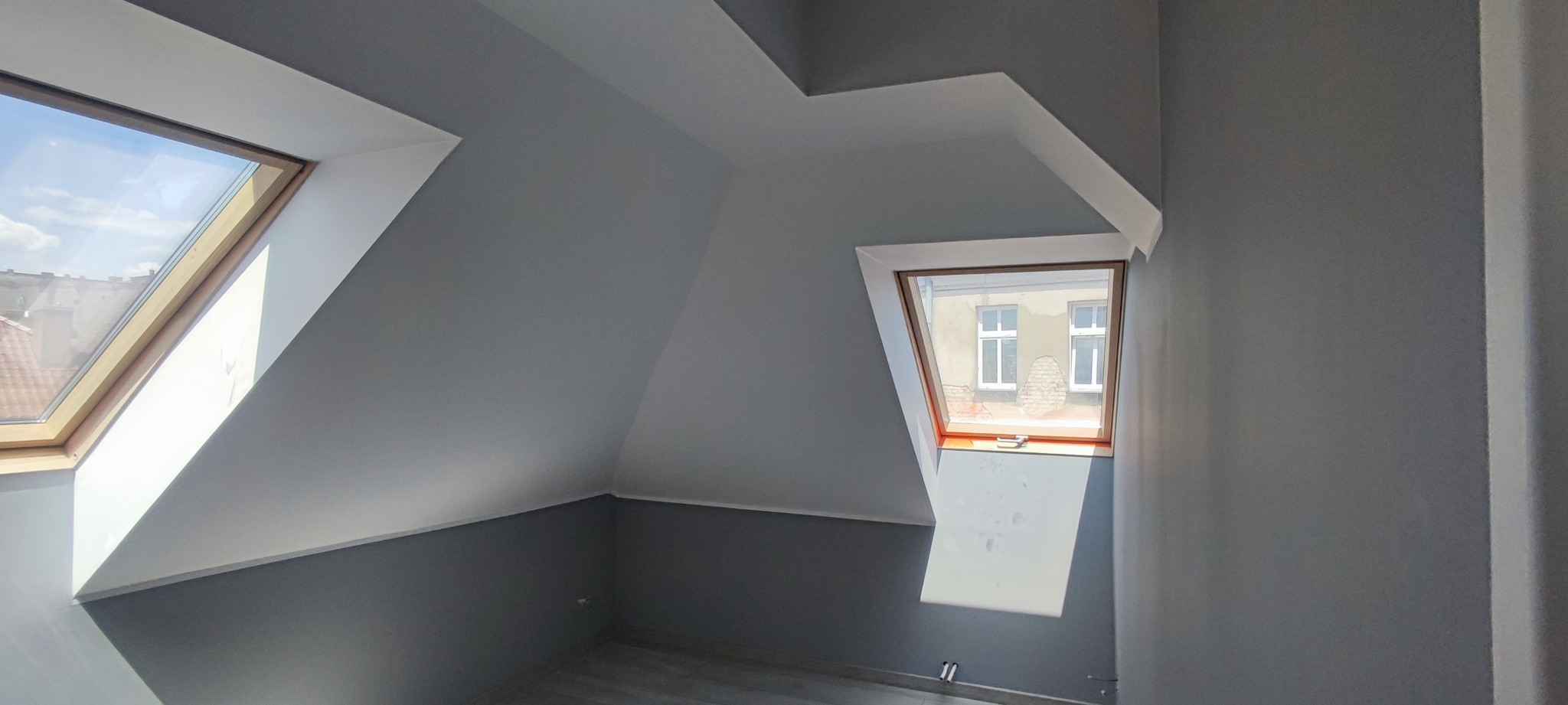 Pomalowane na szaro poddasze z dwoma oknami dachowymi, widok na budynek z cegły za oknem.