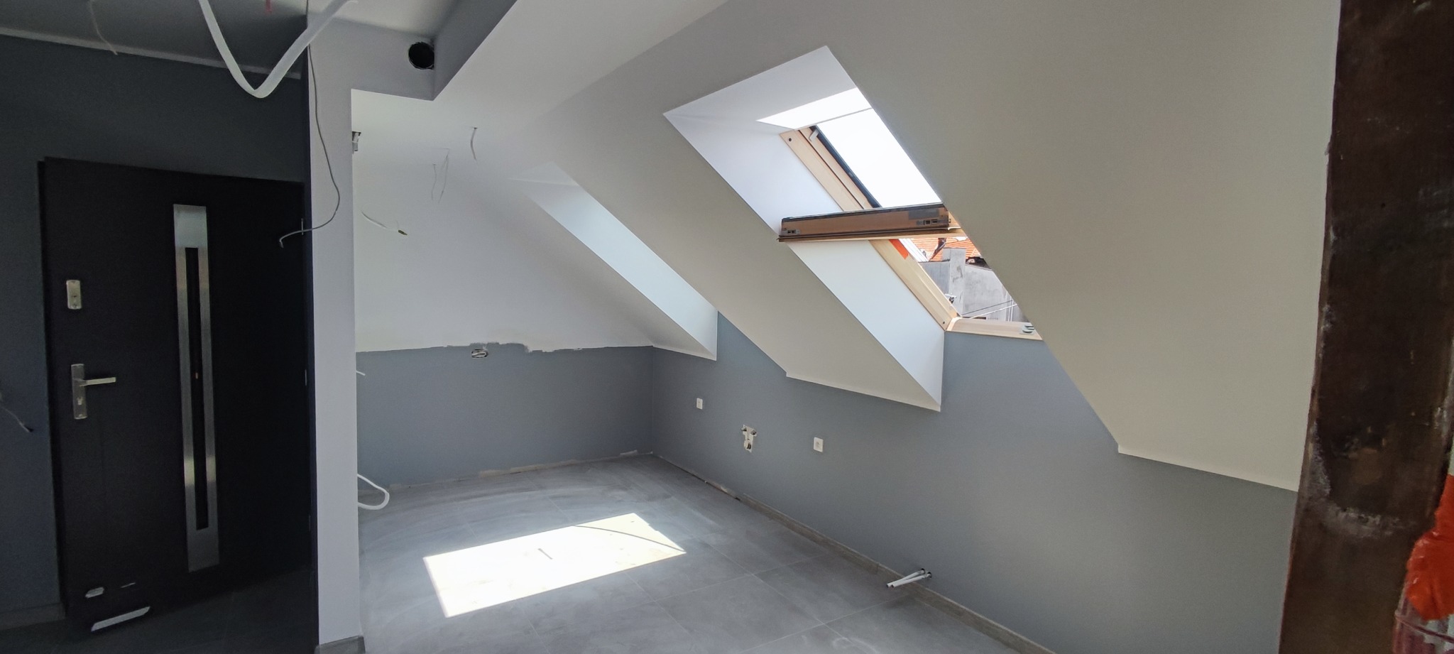 Poddasze po remoncie z szarymi ścianami, dwoma oknami dachowymi i ciemnymi drzwiami wejściowymi, widoczne niedociągnięcia w postaci niezakrytych przewodów elektrycznych i śladów po szpachlowaniu.