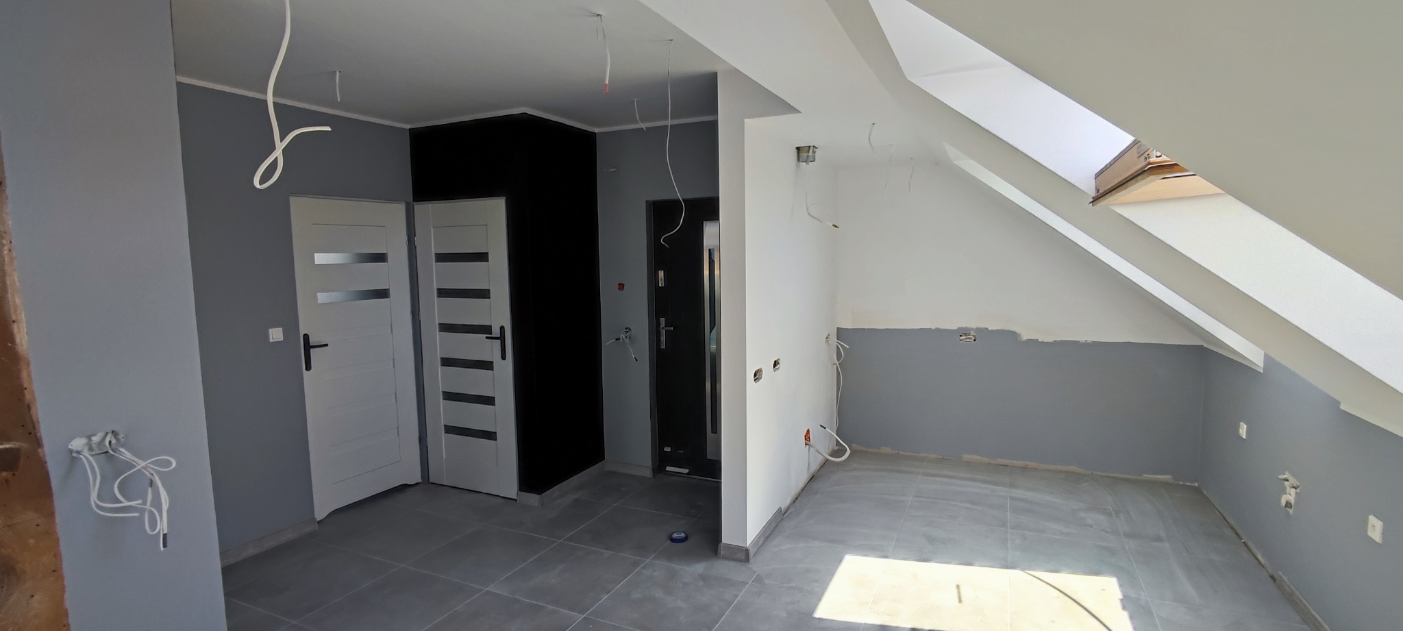Poddasze w trakcie wykończenia: szare ściany, białe skosy, dwie pary drzwi z widocznymi przewodami elektrycznymi, szare płytki podłogowe i czarny fragment ściany.