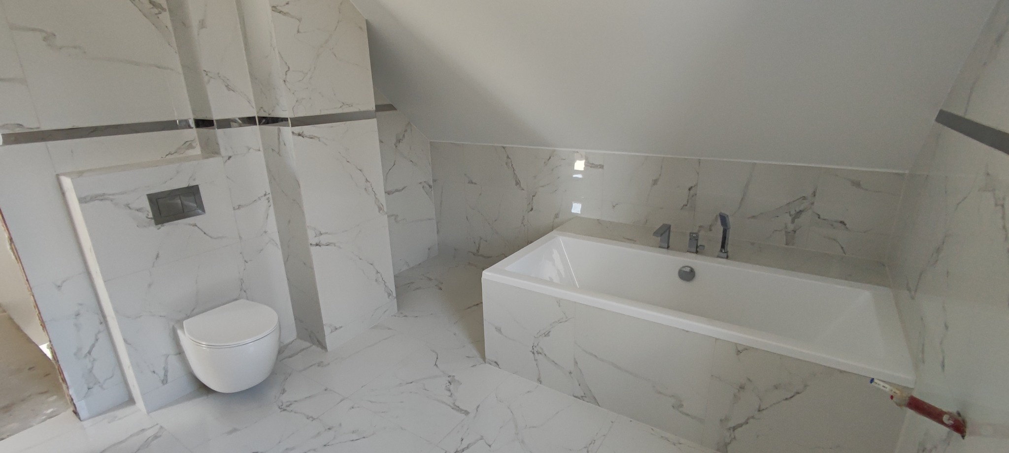 Wykończona łazienka na poddaszu z białą armaturą i marmurowymi płytkami, widoczny fragment pędzla malarskiego przy wannie.