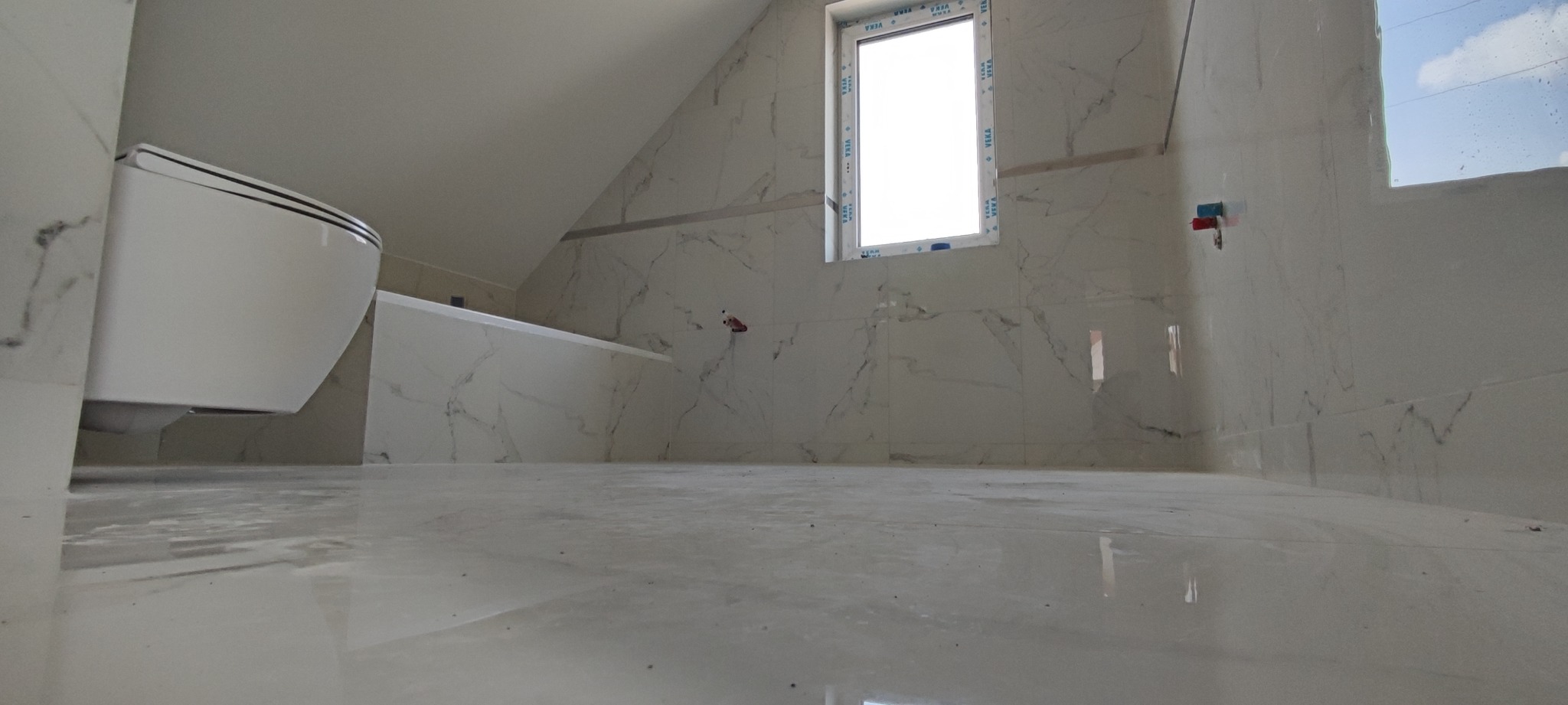 Wykończona łazienka na poddaszu z białymi marmurkowymi płytkami na ścianach i podłodze, wisząca toaleta i okno z białymi ramami.