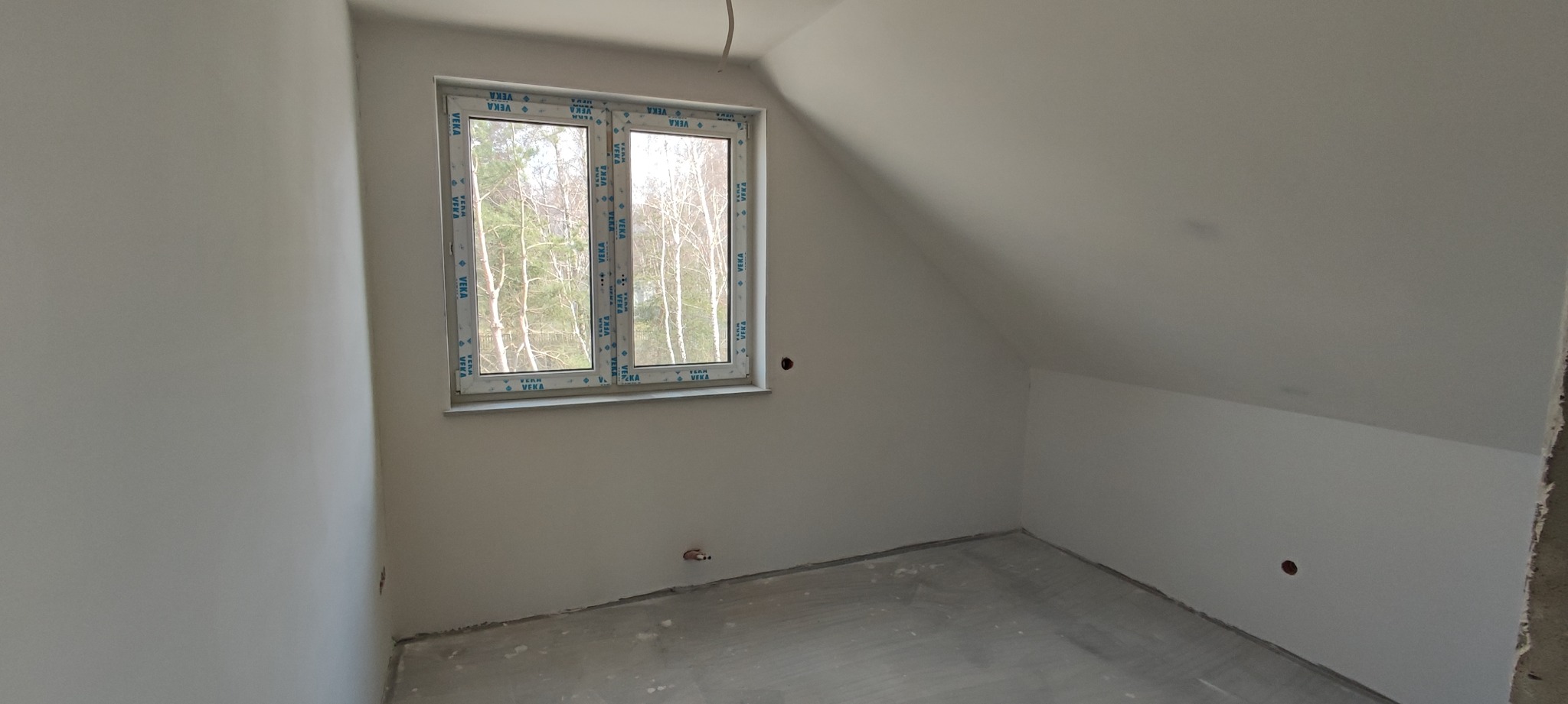 Poddasze w stanie deweloperskim z zamontowanym oknem PCV z widokiem na drzewa, ściany pomalowane na biało z widocznymi otworami instalacyjnymi, surowa wylewka na podłodze.