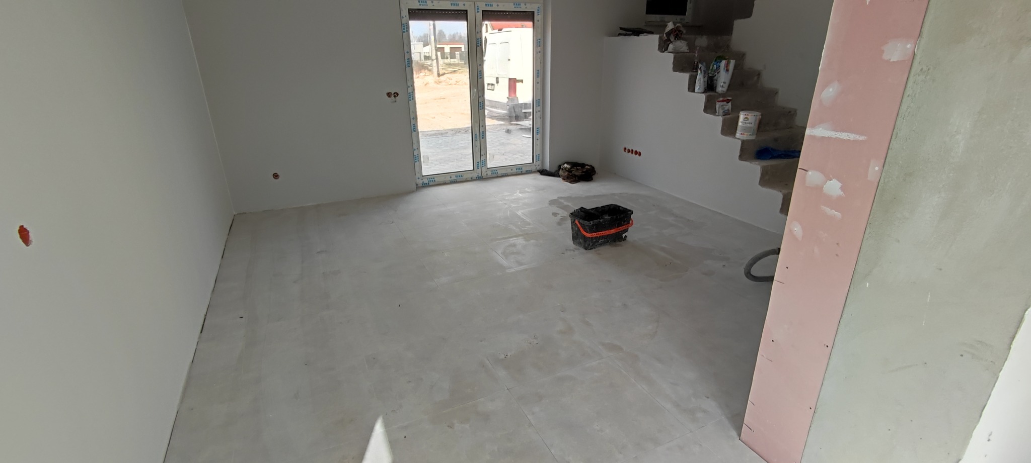 Wnętrze w stanie deweloperskim z przygotowanym podłożem pod posadzkę, widoczne ślady po szlifowaniu, wiadro i odkurzacz przemysłowy, schody w surowym stanie, okno balkonowe z widokiem na budowę.