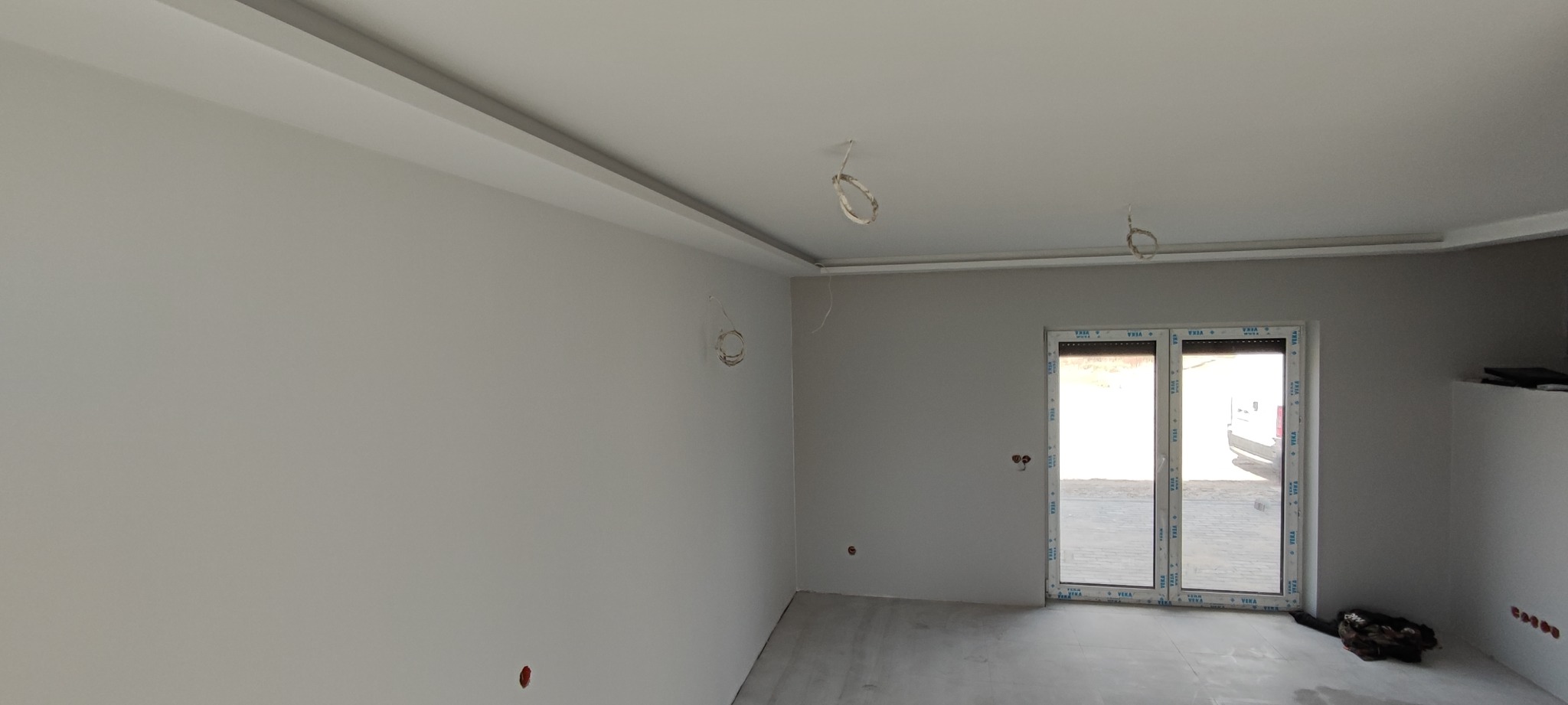 Wnętrze po malowaniu ścian na szaro, widoczne okno PCV z roletami, wystające kable ze ścian i sufitu, oraz listwa dekoracyjna pod sufitem.