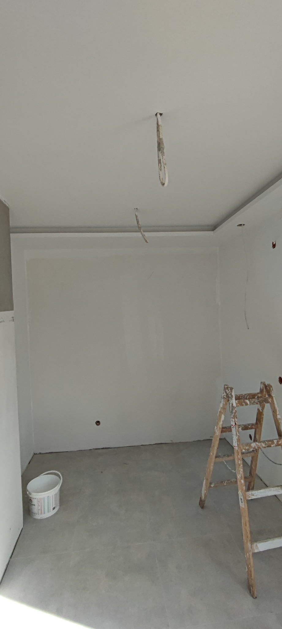 Wnętrze pokoju w trakcie prac wykończeniowych, widoczne gładkie, białe ściany, sufit z dekoracyjną listwą, wystające kable elektryczne, drabina malarska i wiadro z farbą.