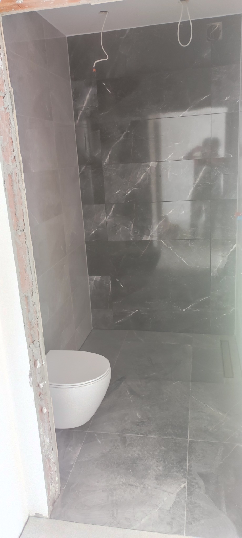 Nowoczesna łazienka w trakcie wykańczania z szarymi płytkami imitującymi marmur, widoczne podłączenie elektryczne i biała miska WC, surowy stan obramowania drzwi.