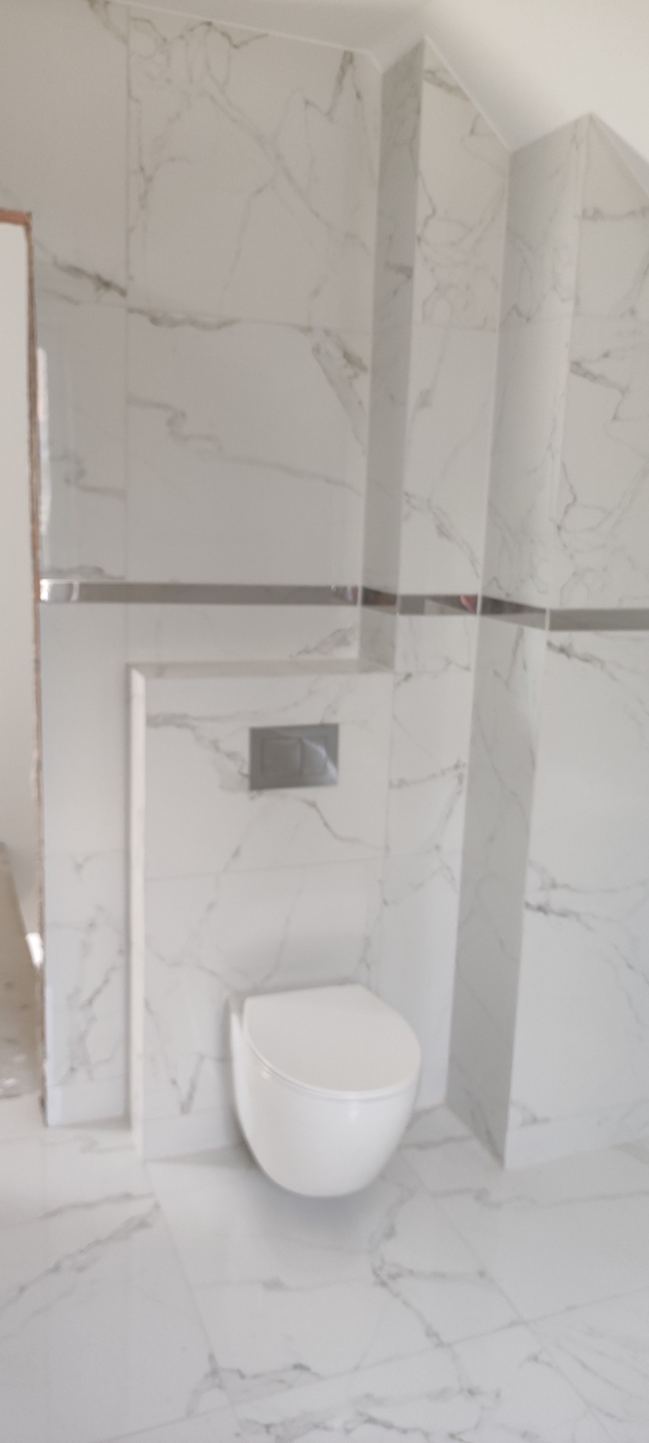 Biała wisząca toaleta na tle ściany i podłogi wyłożonej płytkami imitującymi marmur, z widocznym przyciskiem spłuczki w kolorze srebrnym.