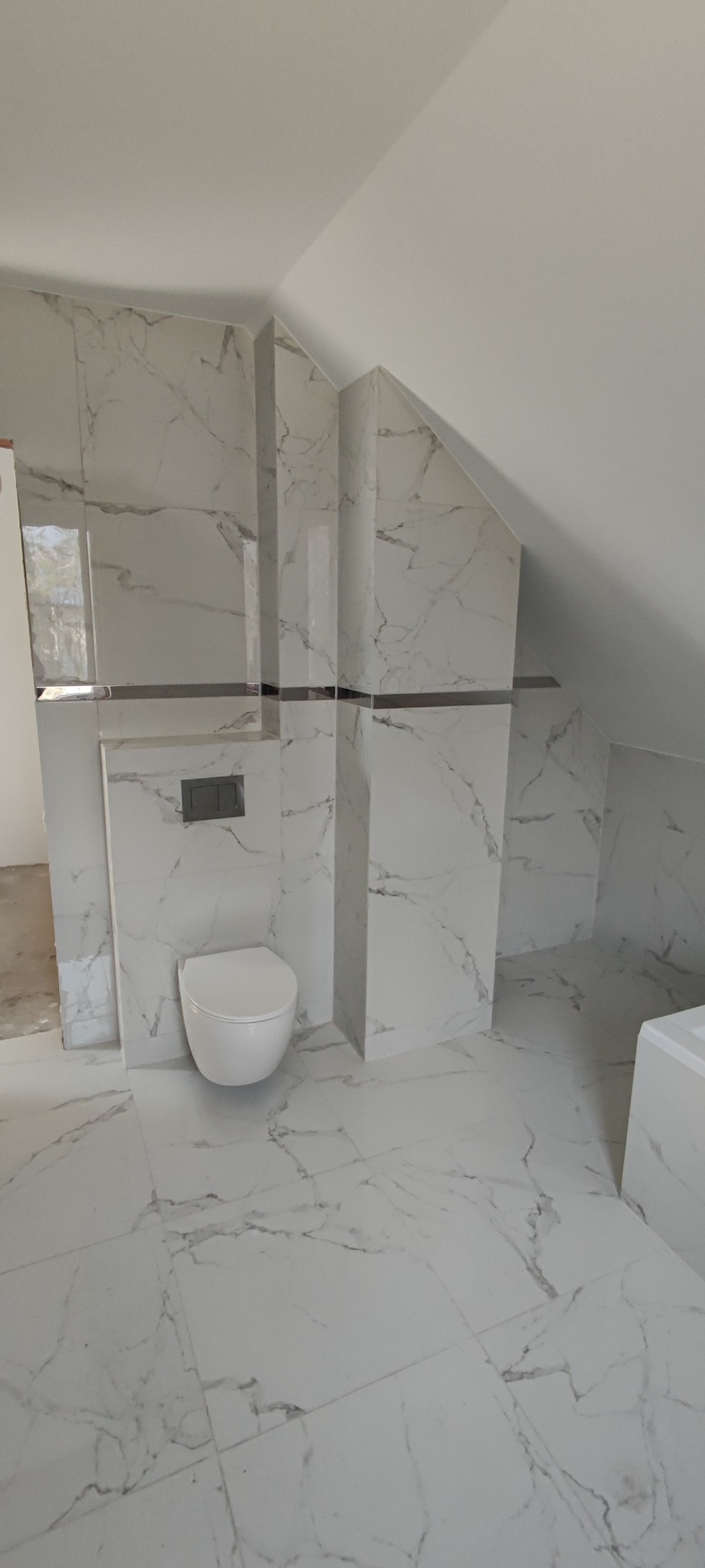 Wykończona łazienka na poddaszu z białą armaturą i płytkami imitującymi marmur, z czarnymi akcentami w postaci listew dekoracyjnych.