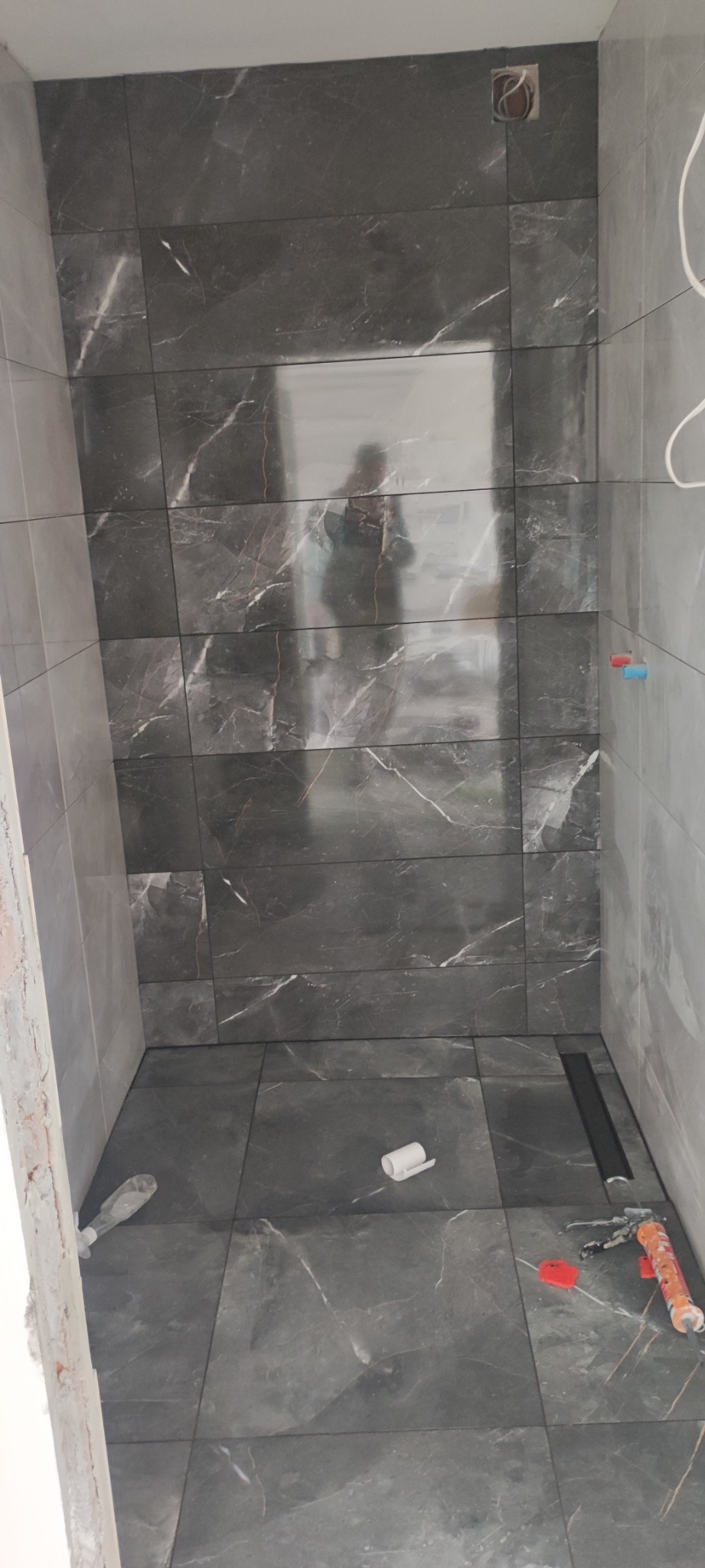 Wykończona łazienka z ciemnymi, marmurkowymi płytkami, widoczny odpływ liniowy, silikon i poziomica na podłodze, oraz odbicie osoby fotografującej w płytkach na ścianie.