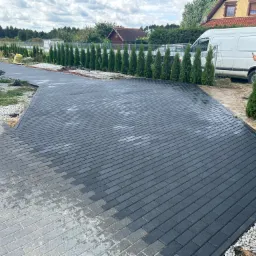 Częściowo ułożona, ciemnoszara kostka brukowa na podjeździe, widoczny biały bus i pracownik z telefonem w tle, obrzeża wykończone białym żwirem i tujami.