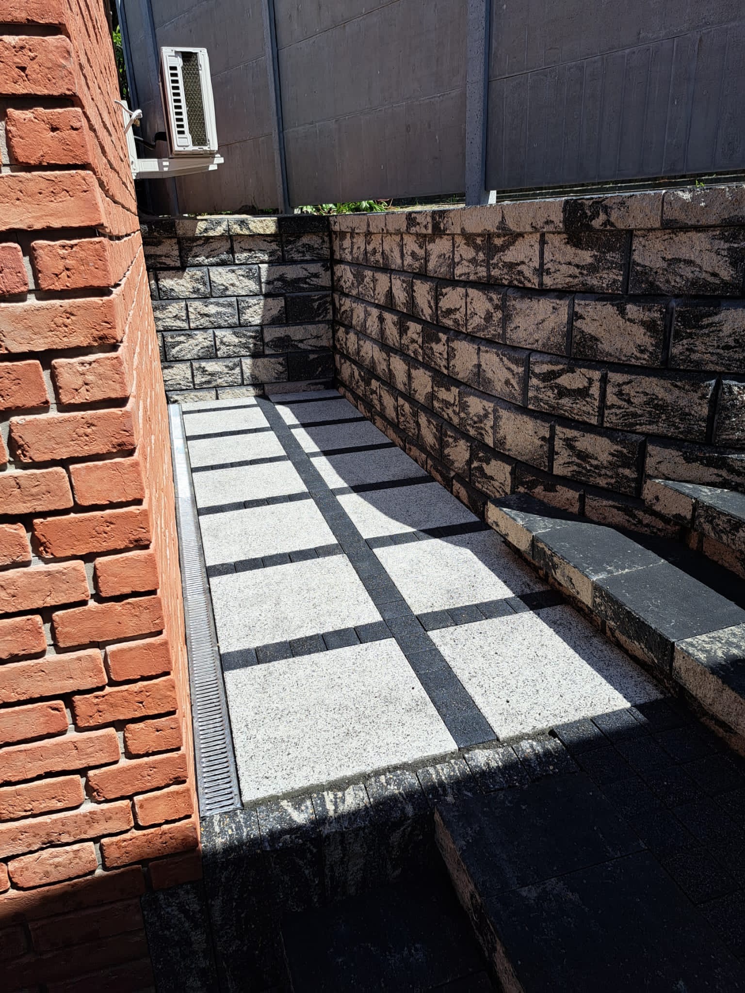 Wykończone patio z jasnej kostki z ciemnymi pasami, otoczone murkiem z ciemnej kostki i ceglaną ścianą, widoczny odpływ liniowy.