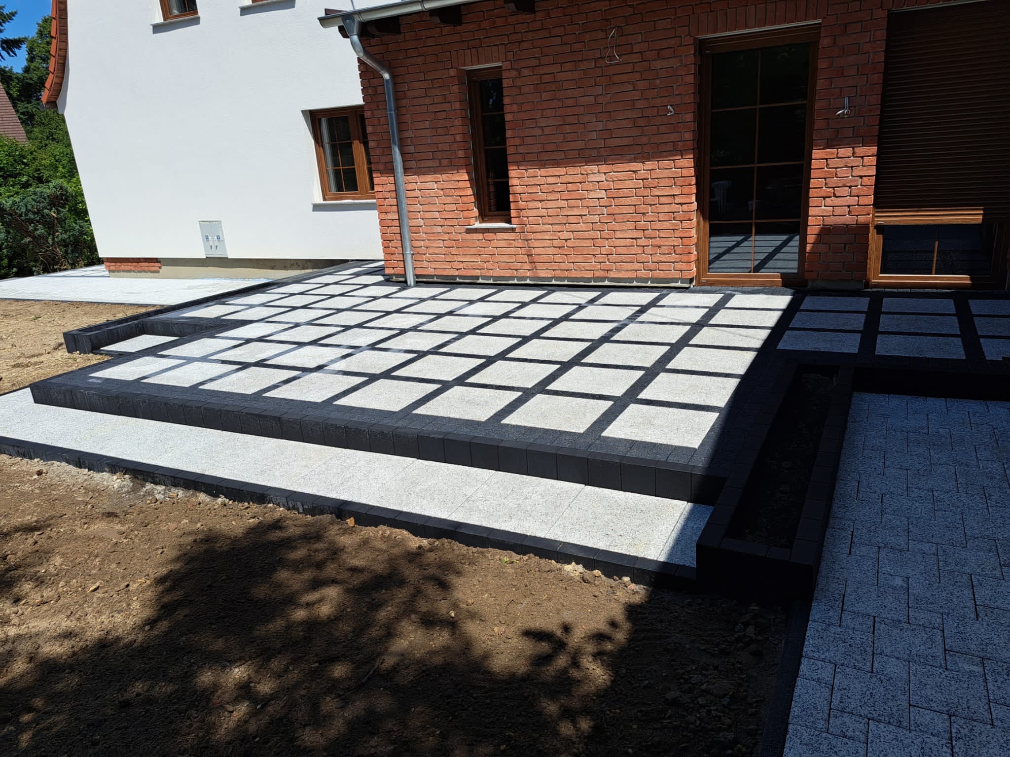 Jasnoszara kostka ułożona w kwadraty z czarnymi obramowaniami, tworząca taras z dwustopniowymi schodami przy budynku z cegły i białym murem.