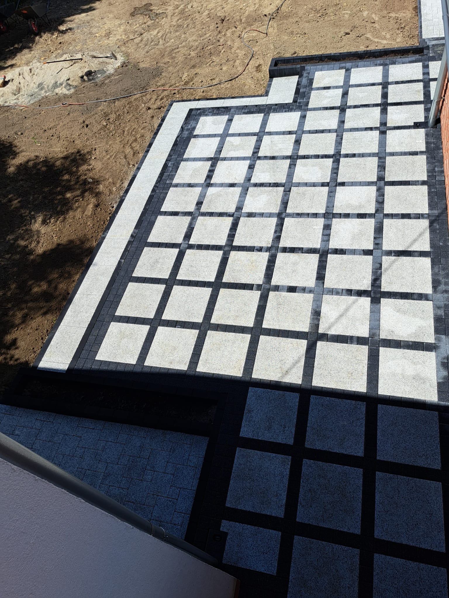 Ułożona kostka granitowa w geometryczny wzór kwadratów, jasnoszarych i ciemnoszarych, z fragmentem ziemi i narzędzi widocznym w tle.