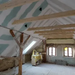 Poddasze w trakcie adaptacji: widoczne drewniane belki stropowe, częściowo zamontowane płyty na ścianach ze śladami szpachlowania, ocieplenie wełną mineralną w przestrzeniach między krokwiami,...