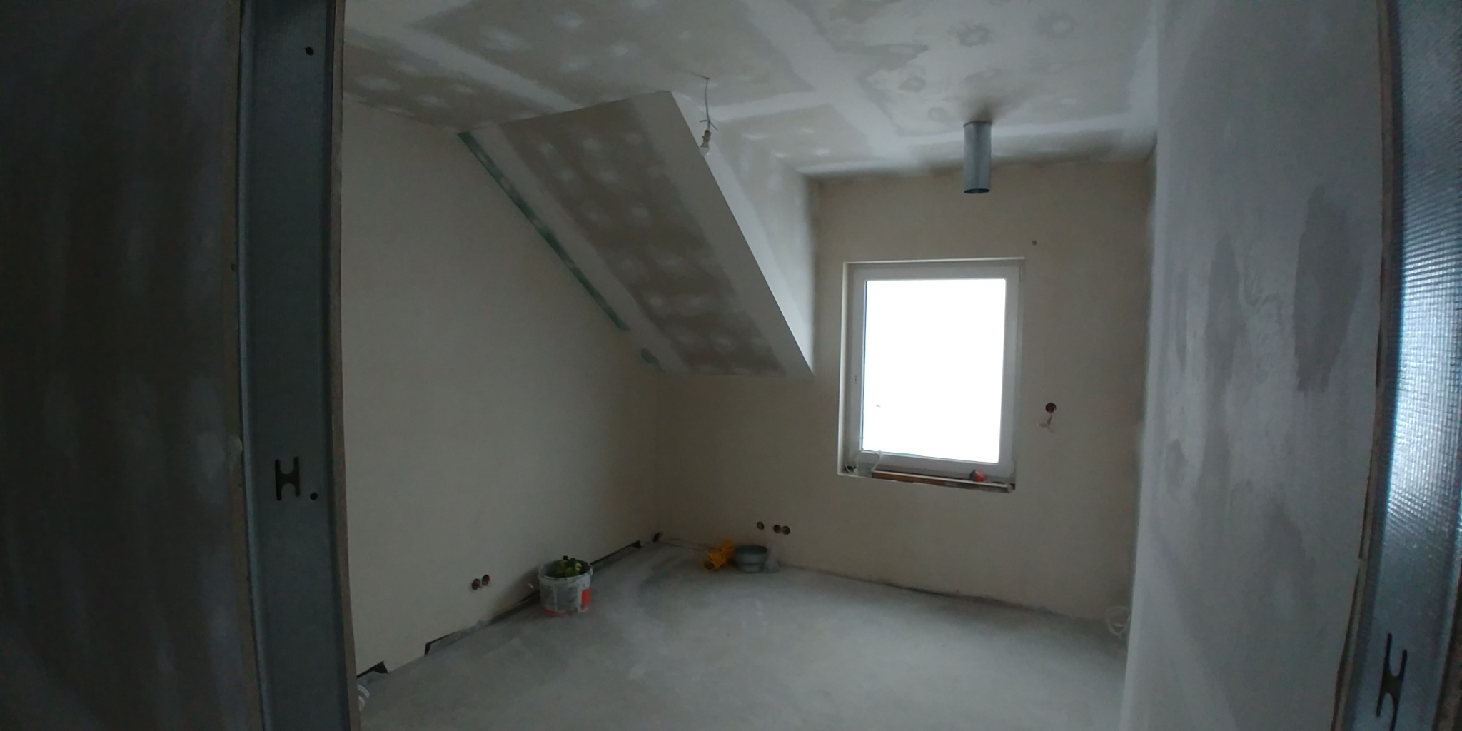 Pokój na poddaszu w trakcie prac wykończeniowych: ściany i sufit po szpachlowaniu, widoczne łączenia płyt, rura wentylacyjna i okno. W rogu stoi wiadro z narzędziami.