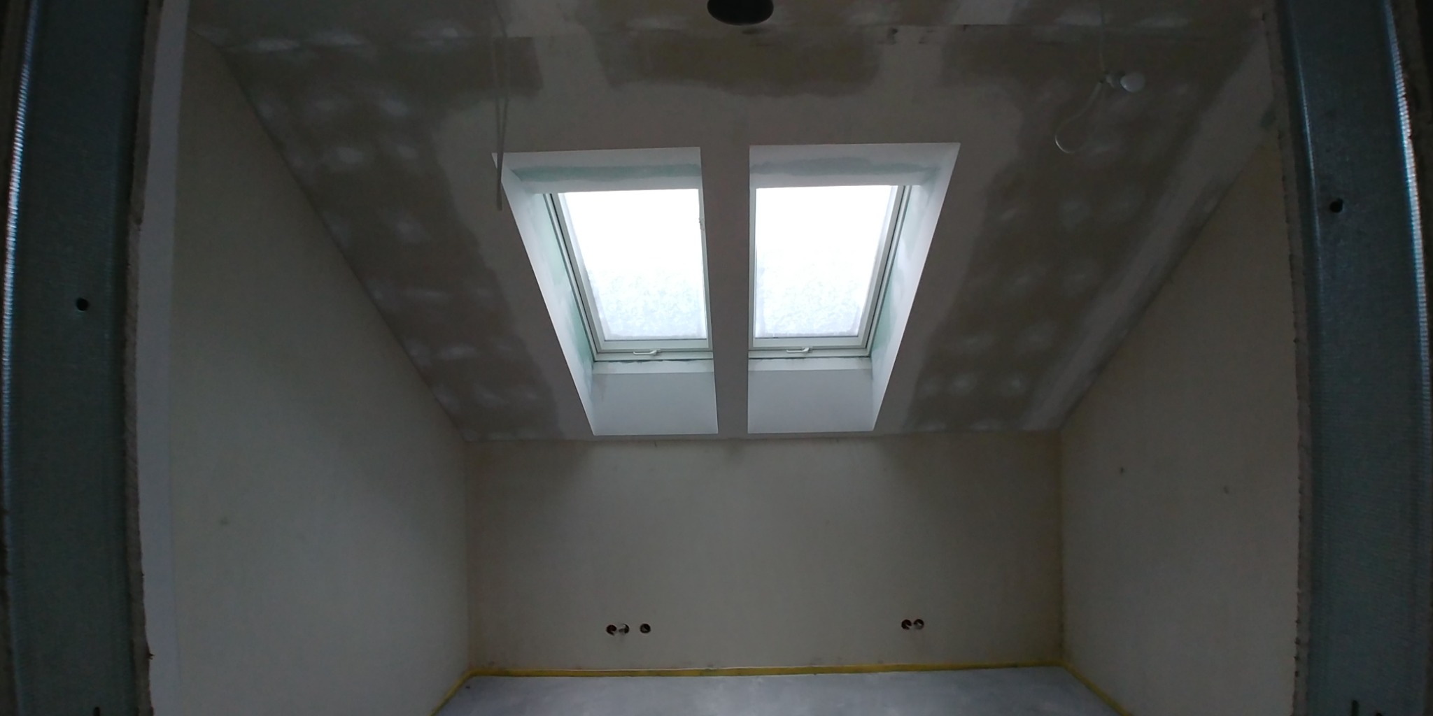 Poddasze w trakcie remontu z dwoma oknami dachowymi, ściany przygotowane do malowania, widoczne profile metalowe na ścianach i suficie, ślady szpachlowania na suficie.