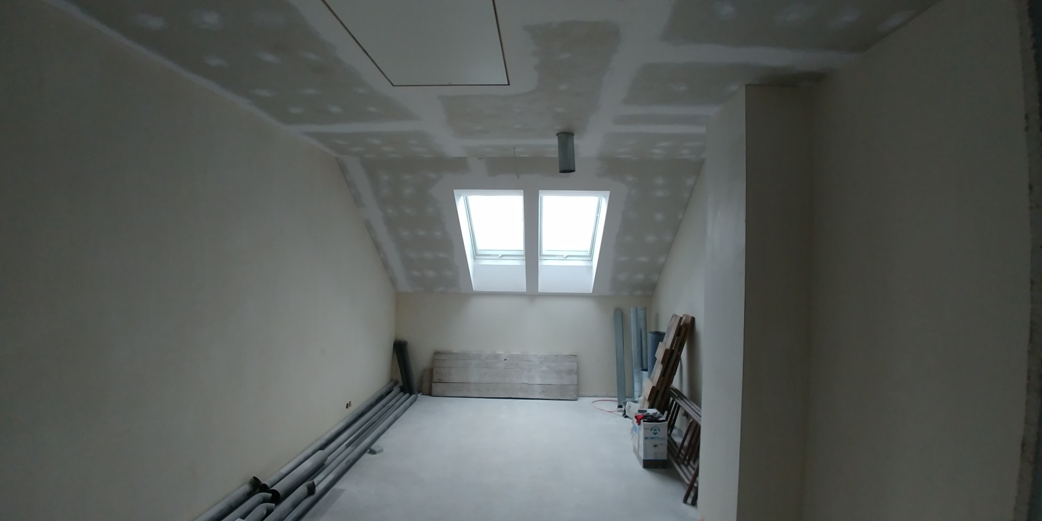 Poddasze w trakcie prac wykończeniowych: ściany przygotowane pod malowanie, widoczne łączenia płyt gipsowo-kartonowych na suficie oraz rury grzewcze przy ścianie, okna dachowe wpuszczające...