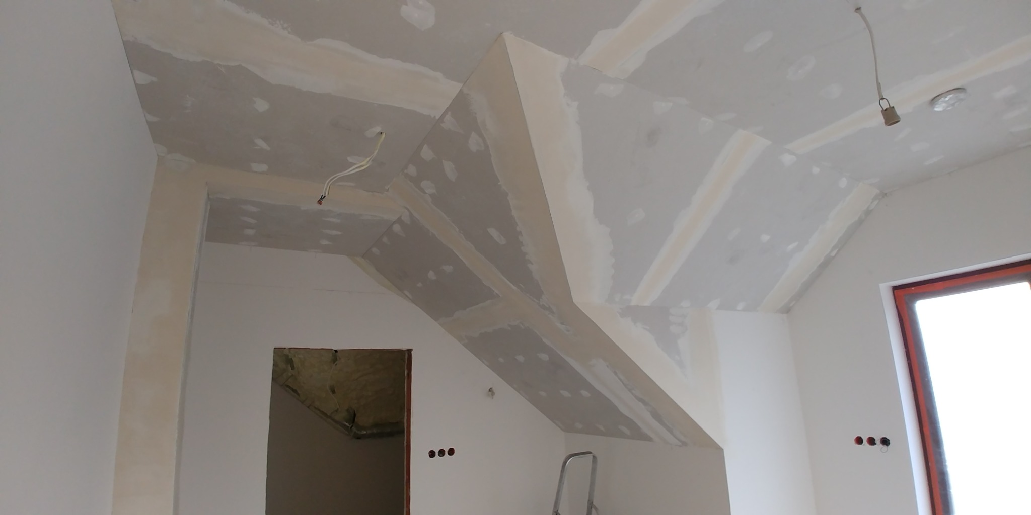 Skośny sufit poddasza z zamontowanymi płytami gipsowo-kartonowymi, widoczne łączenia płyt wymagające szpachlowania, fragment okna i instalacja elektryczna w tle.