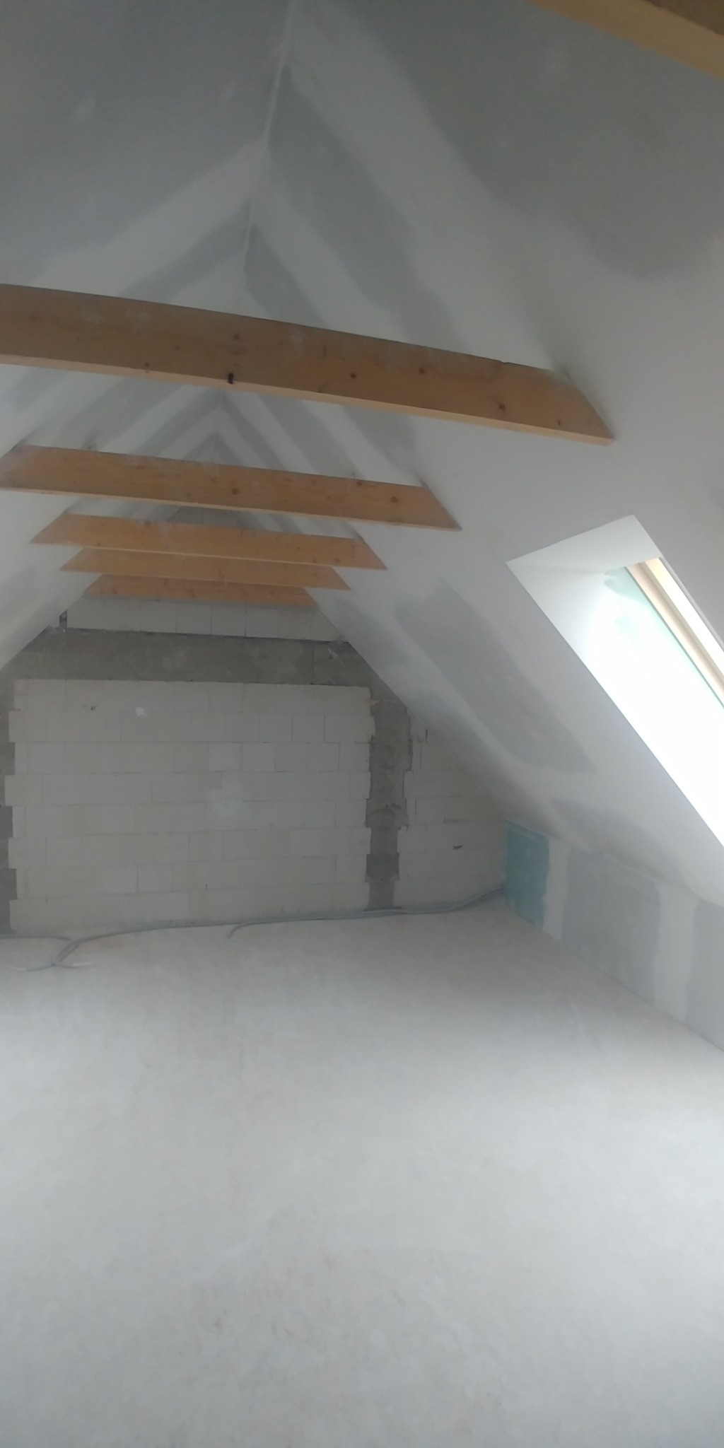 Poddasze w trakcie prac wykończeniowych, widoczne drewniane belki konstrukcyjne, fragment ściany z bloczków, okno dachowe i ściany przygotowane do dalszych prac, prawdopodobnie po szpachlowaniu.