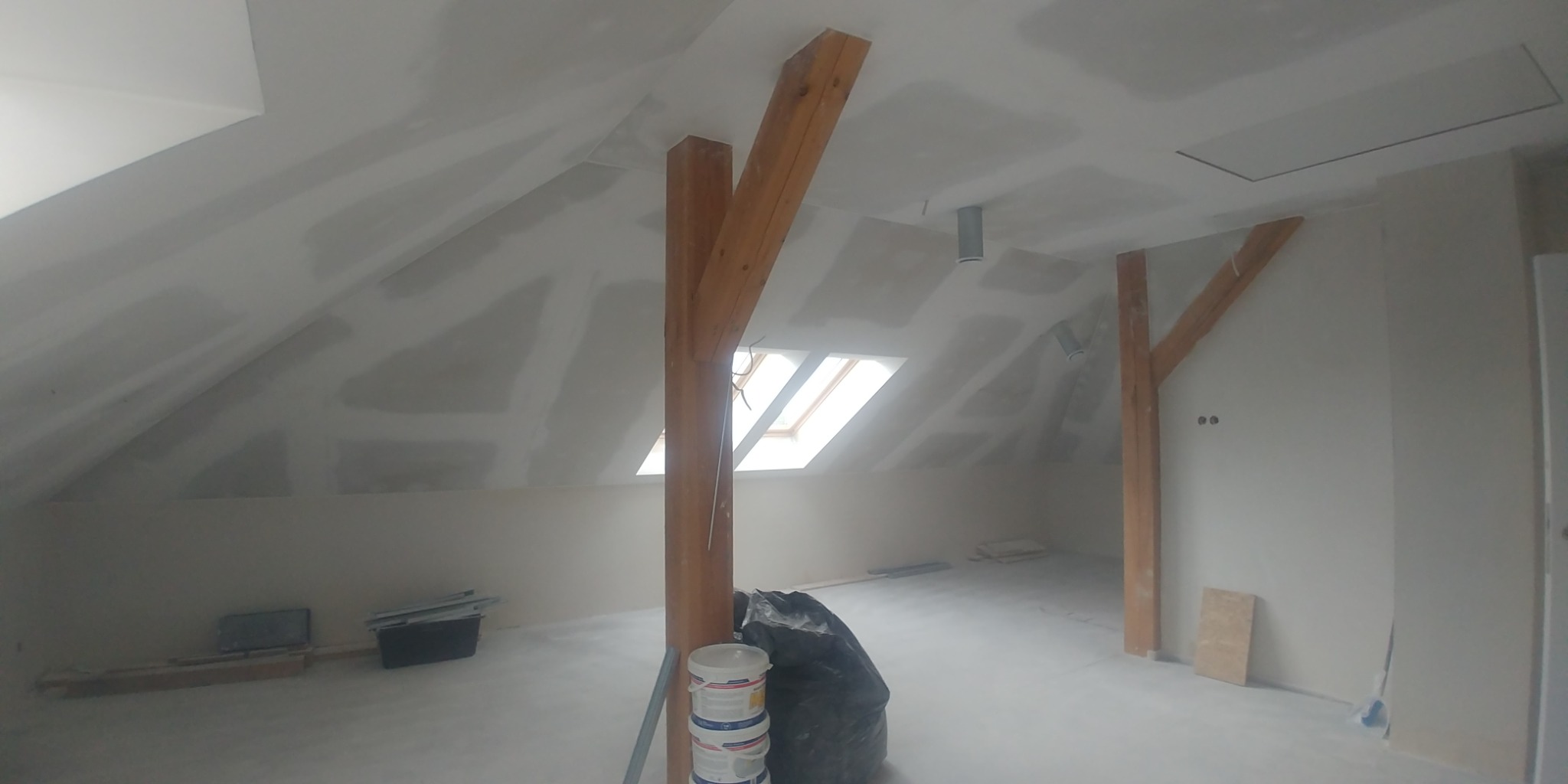 Poddasze w trakcie prac wykończeniowych: drewniane belki konstrukcyjne, okno dachowe, ściany i sufit przygotowane do malowania, widoczne ślady szpachlowania i łączenia płyt, na podłodze pojemniki...