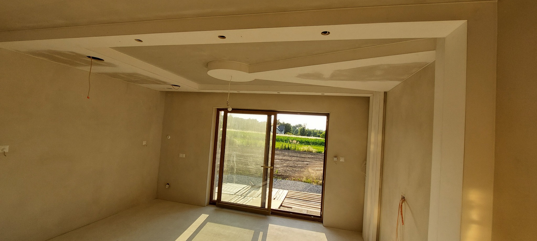 Pokój w trakcie prac wykończeniowych: surowe ściany i sufit z dekoracyjnymi elementami z płyt gipsowo-kartonowych, widoczne otwory na oświetlenie i wystające przewody, duże okno przesuwne...