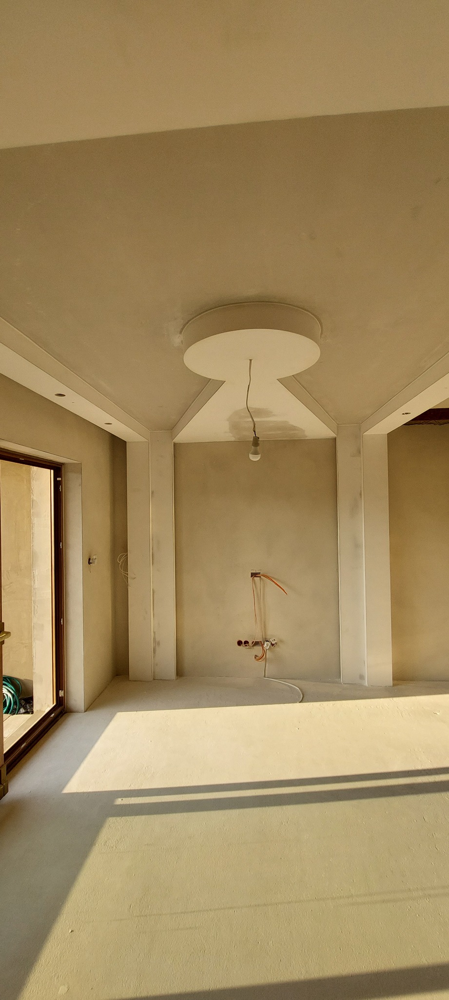 Pokój w stanie deweloperskim z surowymi ścianami, przygotowany do malowania; widoczne instalacje elektryczne i oświetlenie tymczasowe.