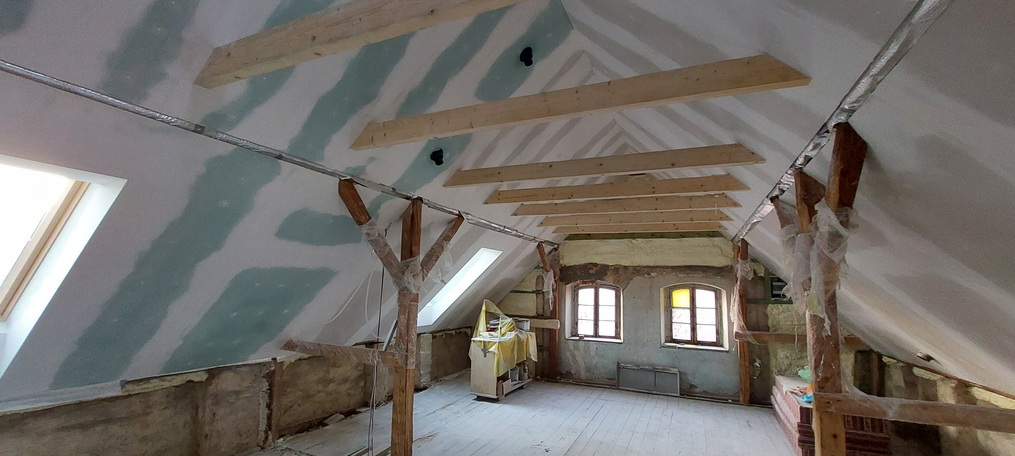 Poddasze w trakcie adaptacji: widoczne drewniane belki stropowe, częściowo zamontowane płyty na ścianach ze śladami szpachlowania, ocieplenie wełną mineralną w przestrzeniach między krokwiami,...