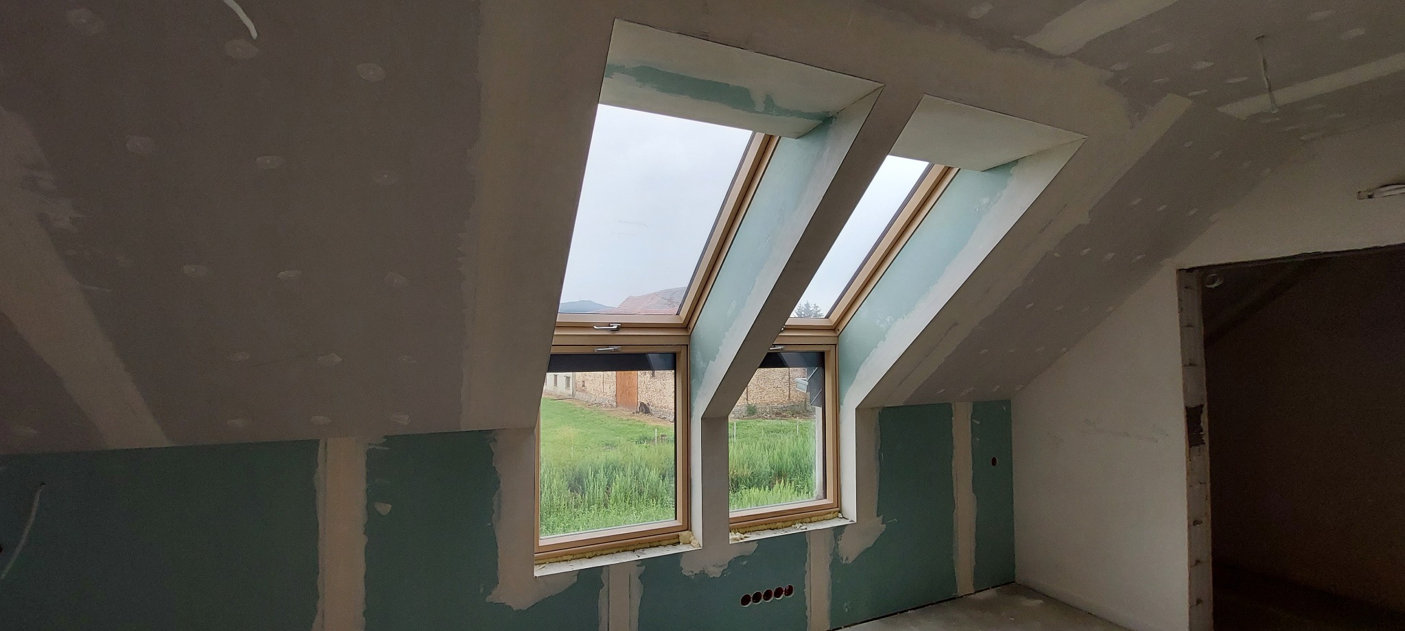 Poddasze w trakcie prac wykończeniowych: zamontowane okna dachowe z widokiem na zieleń, ściany i sufit pokryte płytami gipsowo-kartonowymi, widoczne łączenia płyt i szpachlowanie.