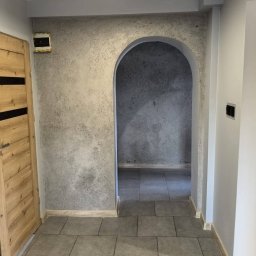 Usługi ogólnobudowlane Dawid Derbich - Wykończony korytarz z łukowatym przejściem i szarą, teksturowaną ścianą. Na podłodze szare płytki. Drewniane drzwi z czarnymi akcentami po lewej stronie.