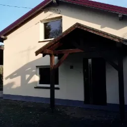 Jasny, otynkowany dom jednorodzinny z drewnianą wiatą nad wejściem i zadaszeniem na boku, widoczny w słoneczny dzień.